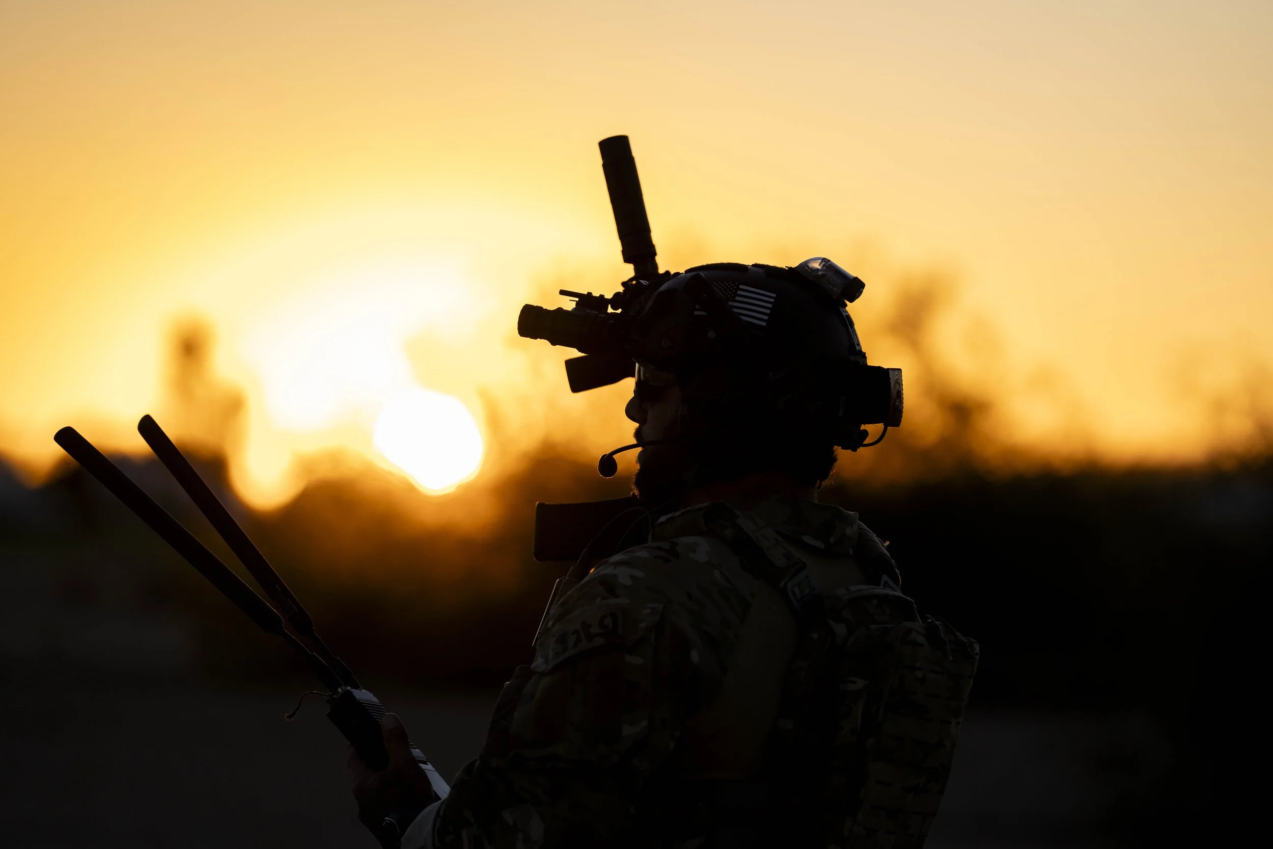 Silhouette of a soldier with military gear and helmet, holding a walkie-talkie, against a setting sun with an orange sky.