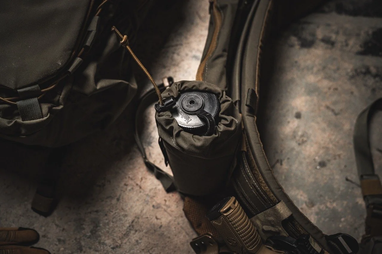 A military-style water bottle with a black lid, attached to a tactical backpack with multiple compartments and straps.