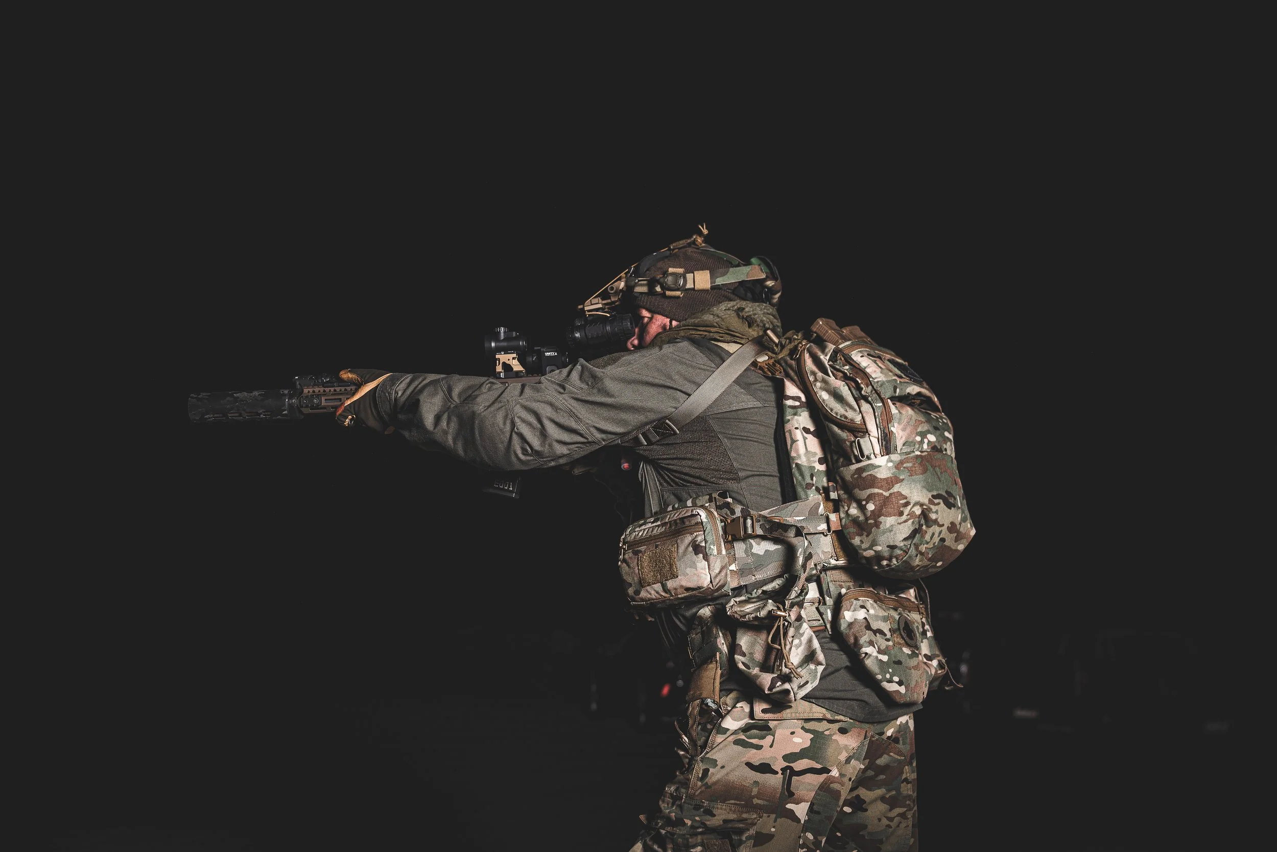 Military soldier aiming rifle in dark environment, wearing camouflage gear and tactical backpack.