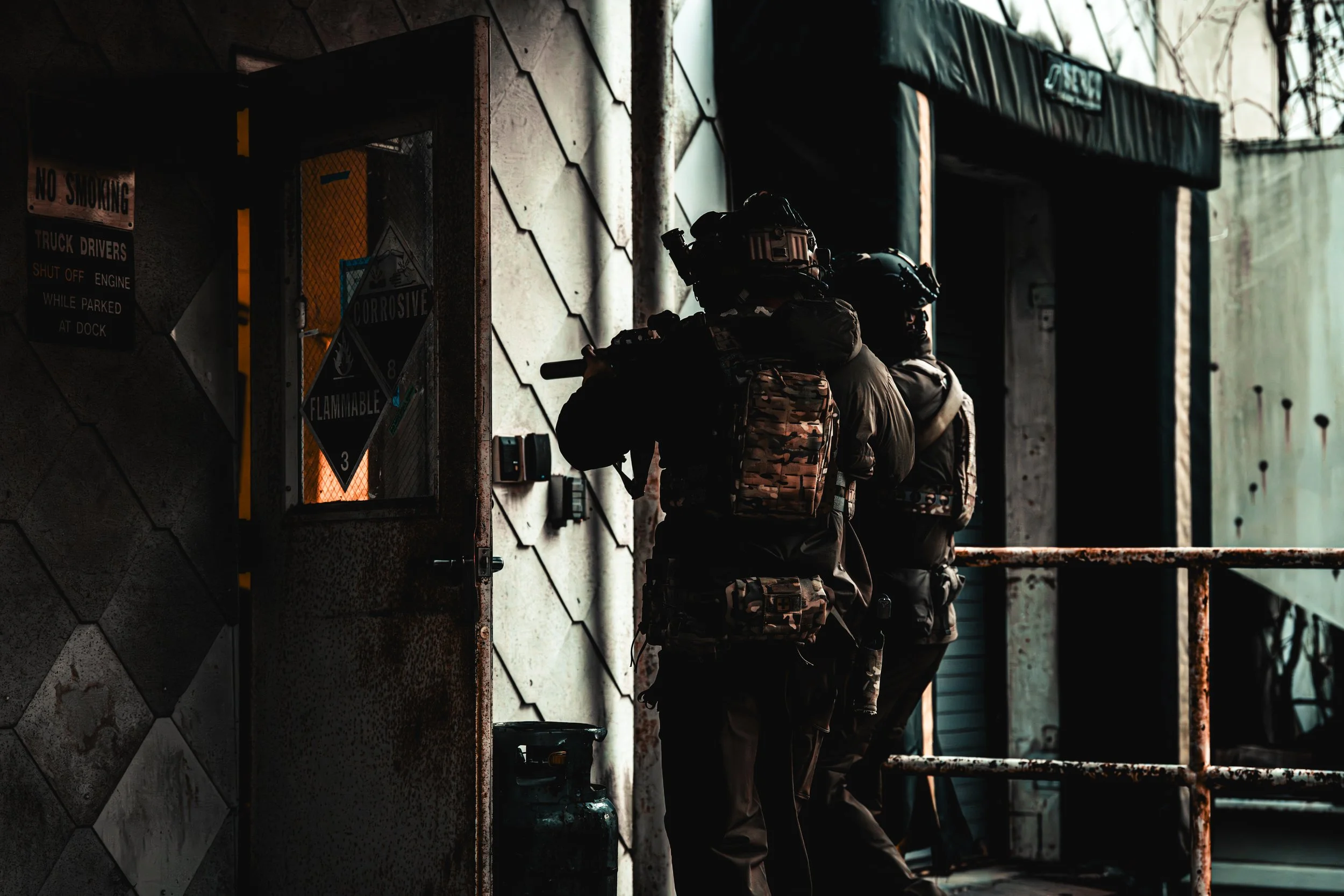 Two armed soldiers in tactical gear moving through a dark, industrial-style door entryway with rusted railing and warning signs on the wall.