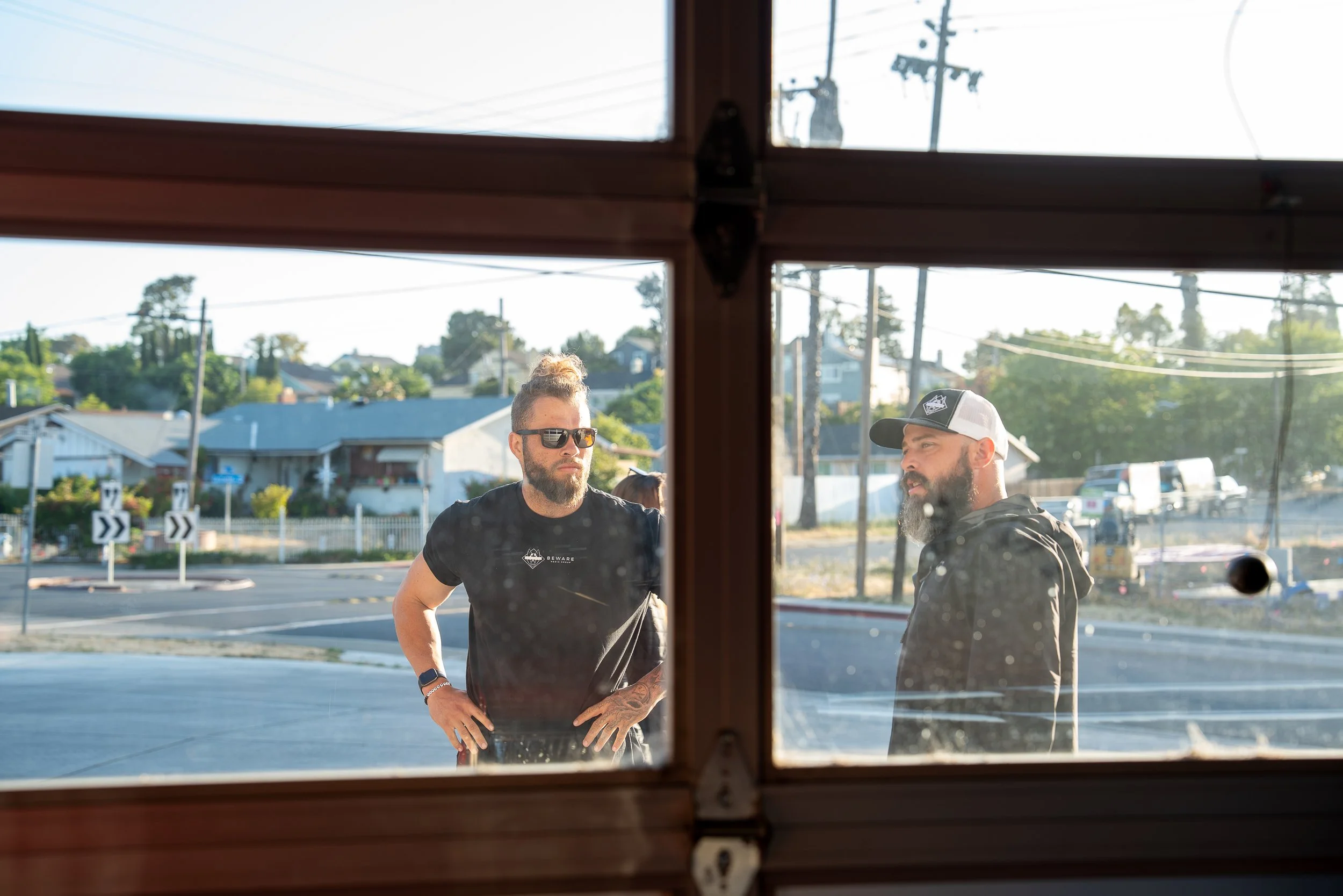 Two men stand outside near a parking lot, seen through a slightly dirty window with a wooden frame. One man has a beard, sunglasses, and an updo hairstyle, while the other has a beard and wears a baseball cap. The background shows residential houses,