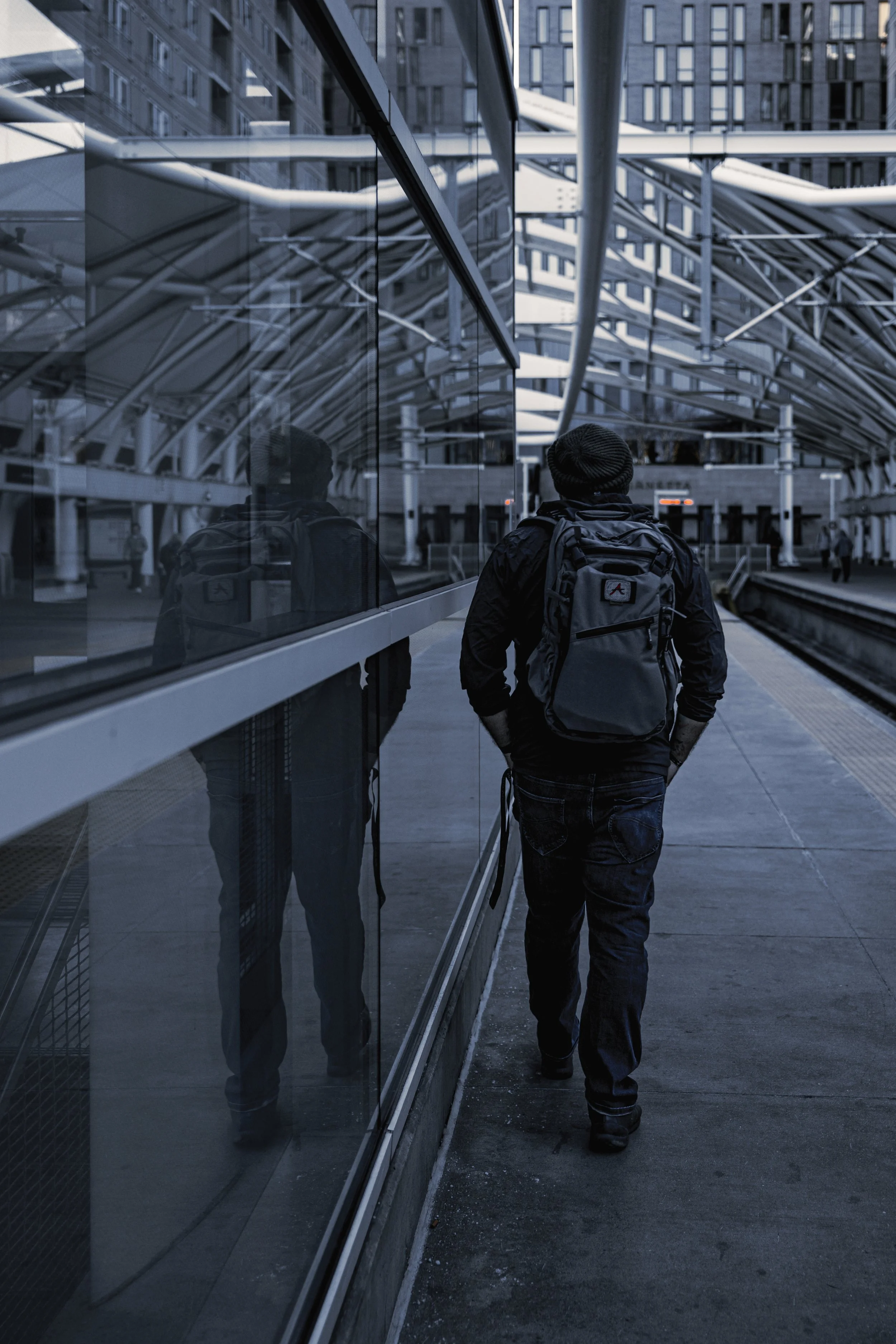A man with a backpack walking along a train station platform beside a glass wall, with city buildings reflected in the glass and visible in the background.
