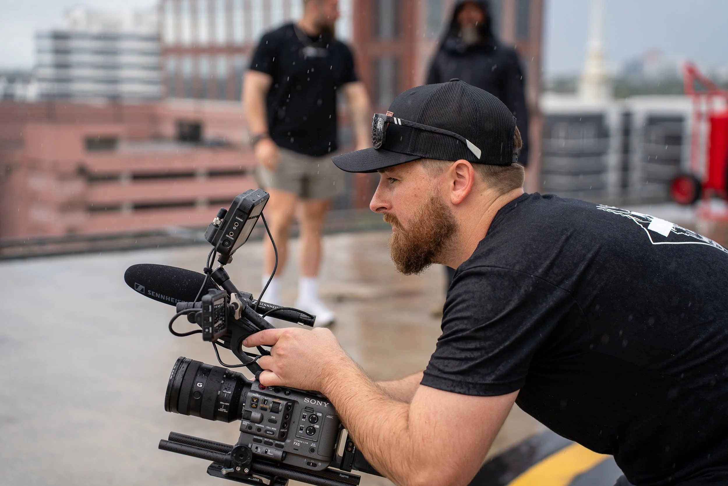 Man recording video with professional camera on rooftop during rain, with two women walking in the background.