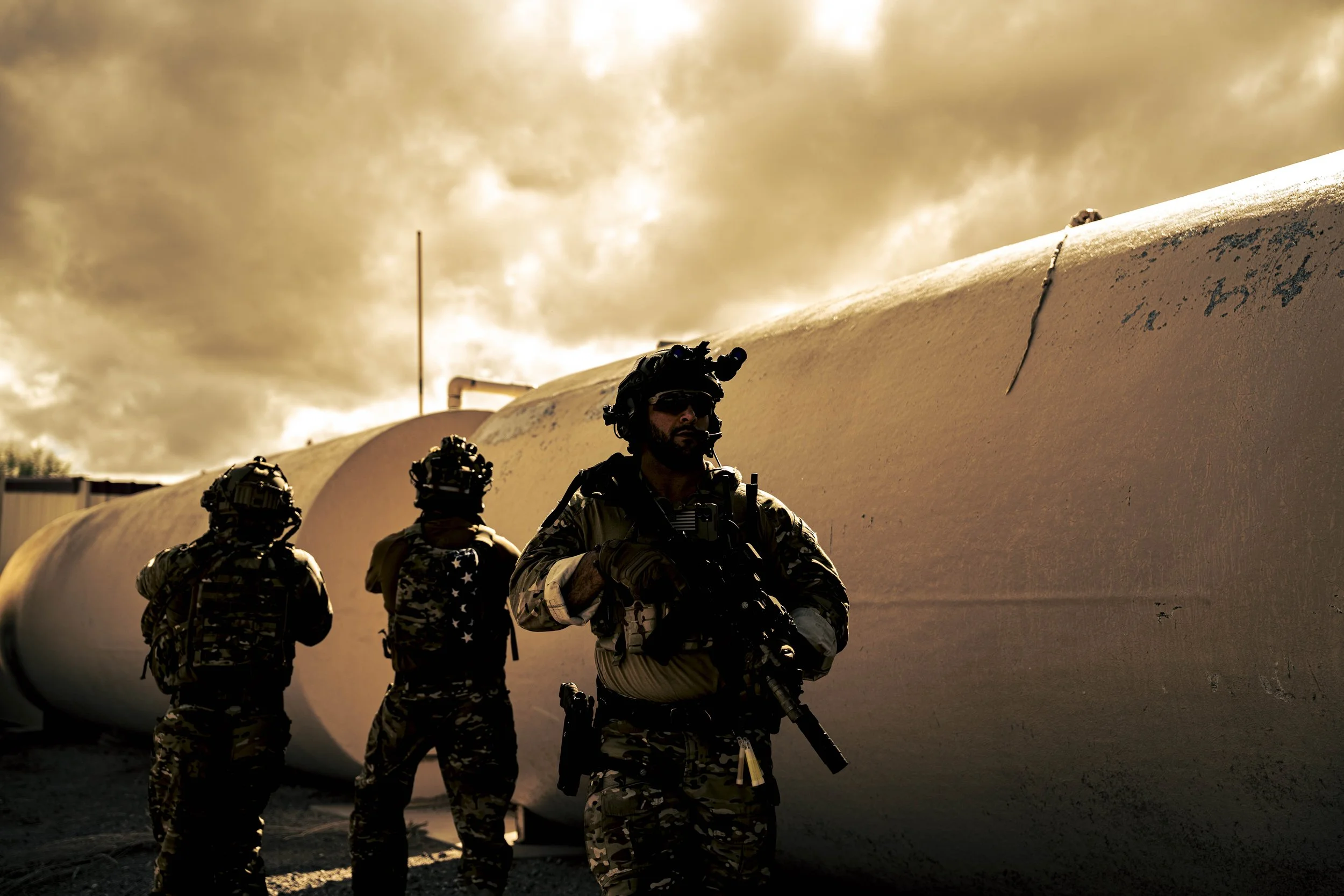 Three soldiers in camouflage uniforms and tactical gear, including helmets and night vision equipment, standing next to a large pipeline under a cloudy sky.