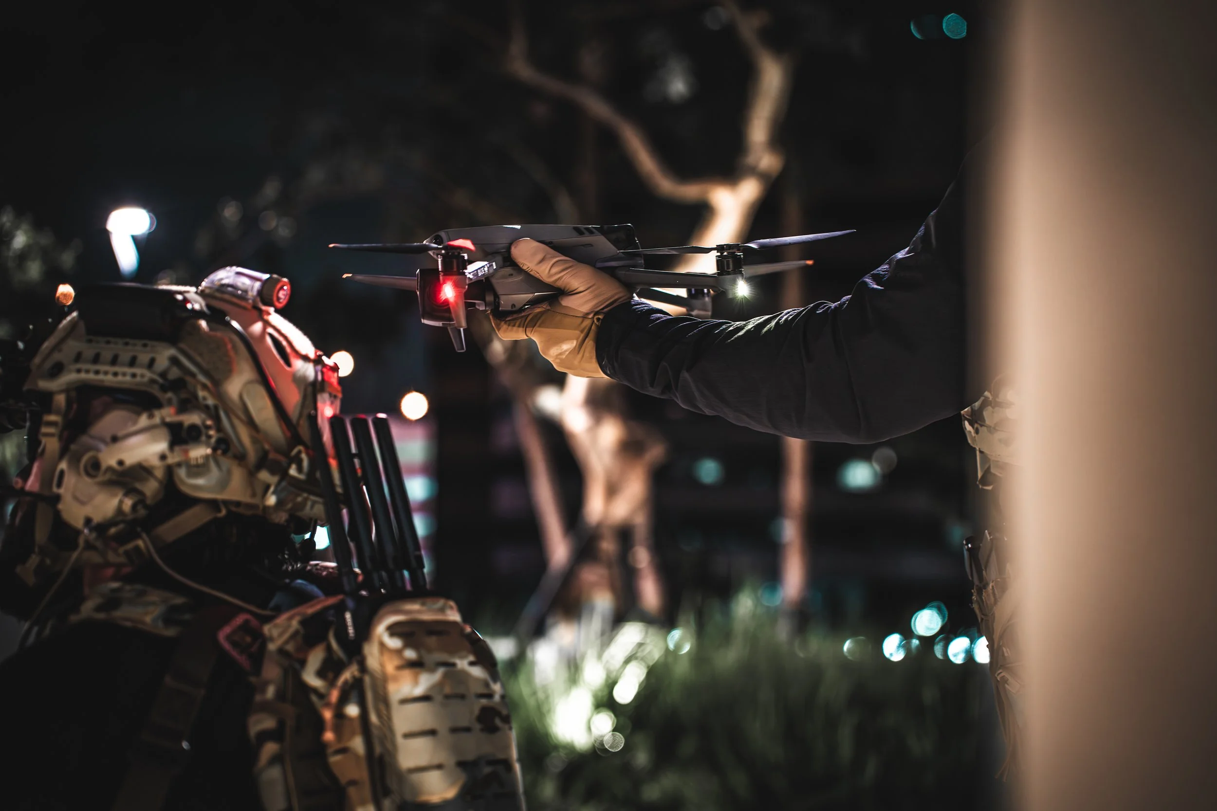 Person holding a drone with a red light illuminated, near a military-style bag at night.