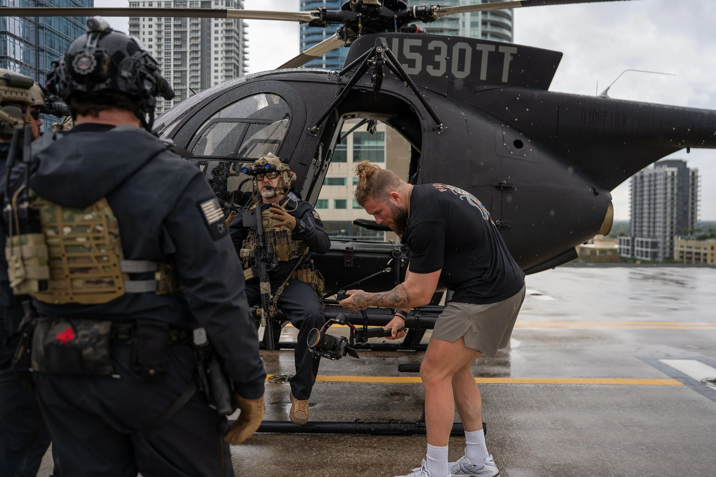 A group of military personnel preparing a black helicopter on an urban rooftop, with tall buildings in the background. One man is sitting inside the helicopter, another is handling equipment, and a third is standing nearby.