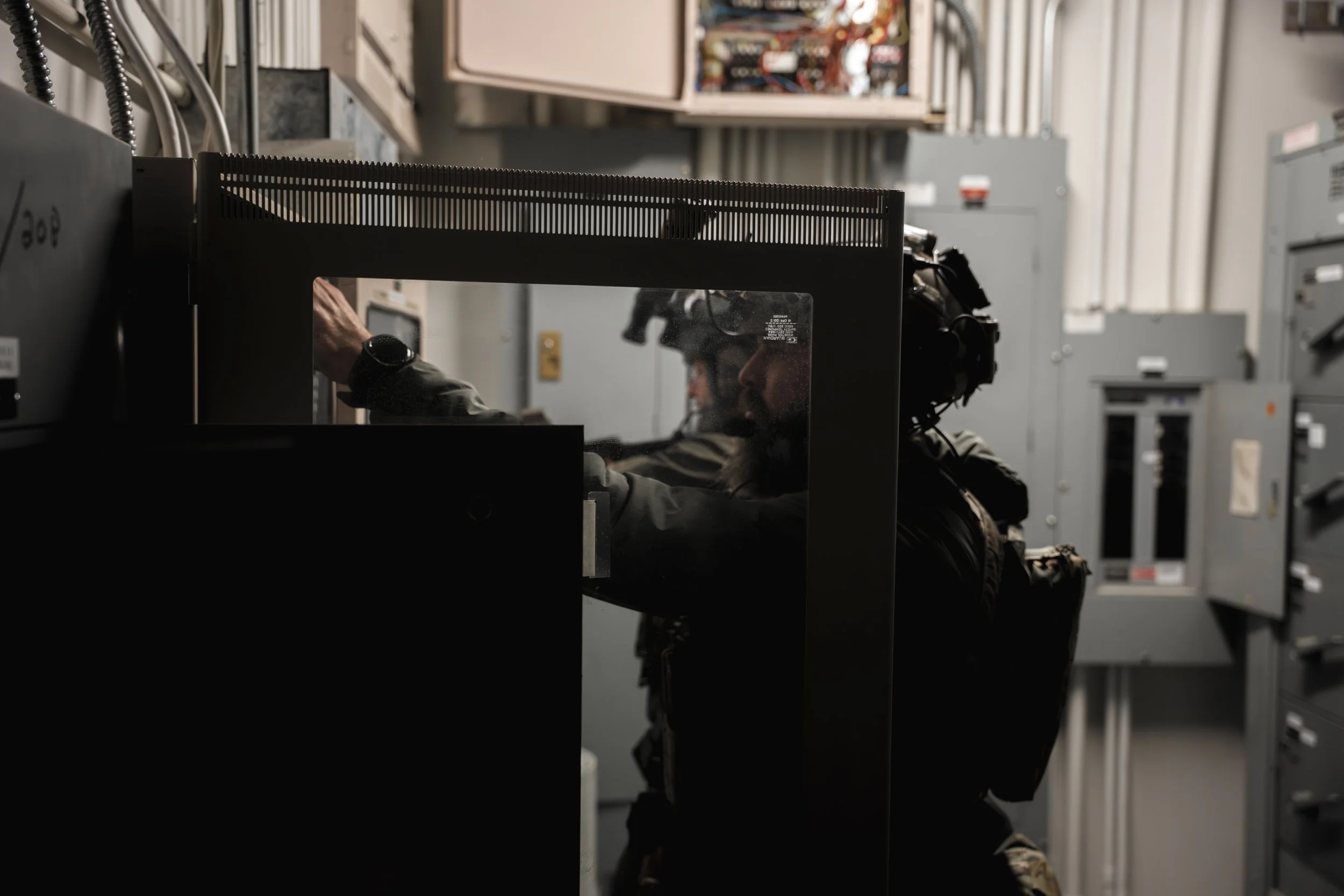 A technician wearing safety glasses and a watch working on electrical panels or equipment inside a utility room with multiple gray electrical boxes.