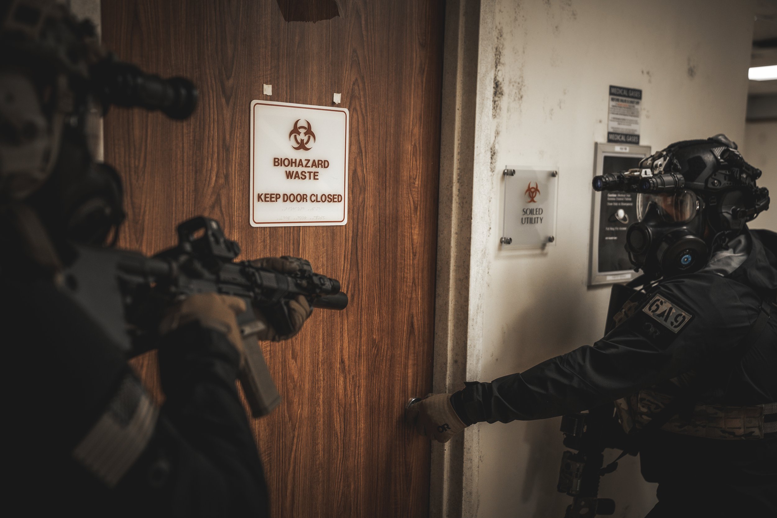 Two individuals in tactical gear and helmets with weapons, closing a door labeled 'Biohazard Waste' in a secure facility.
