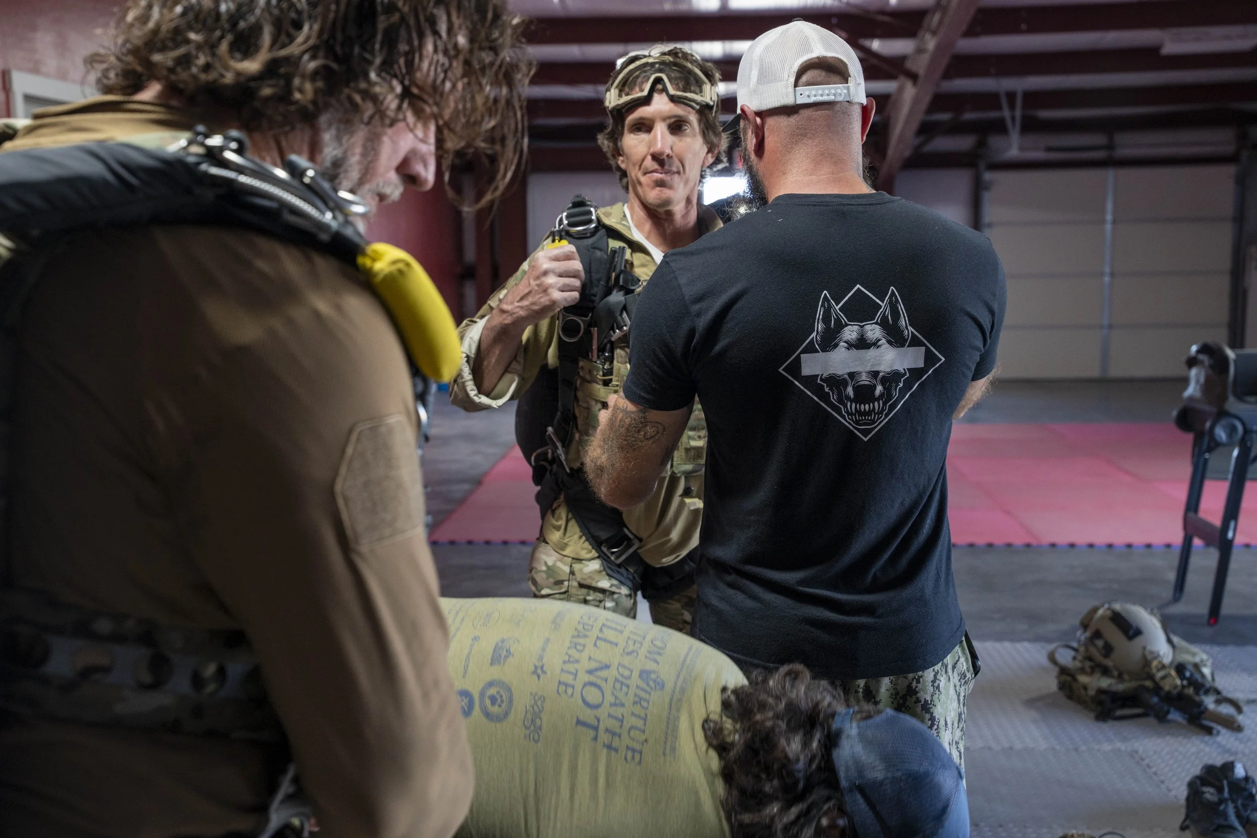 Three men in a gym or training facility, two wearing camouflage gear and a black t-shirt with a wolf logo on the back, the third with curly hair and a banana necklace, engaged in a discussion. Equipment such as a helmet and vest are on the floor.