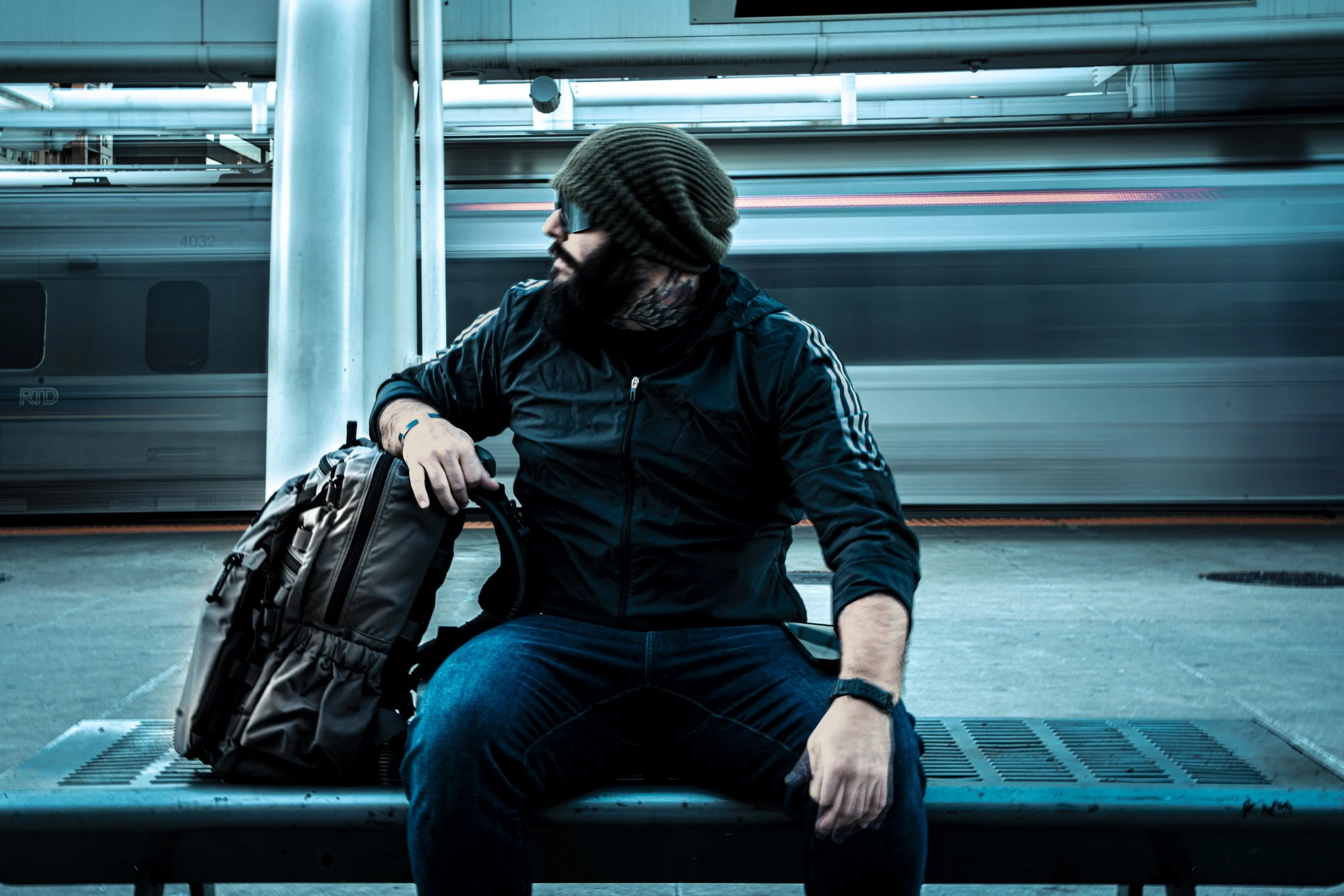 A man in a black jacket, beanie, and sunglasses sitting on a bench at a train station with a blurred train passing in the background.