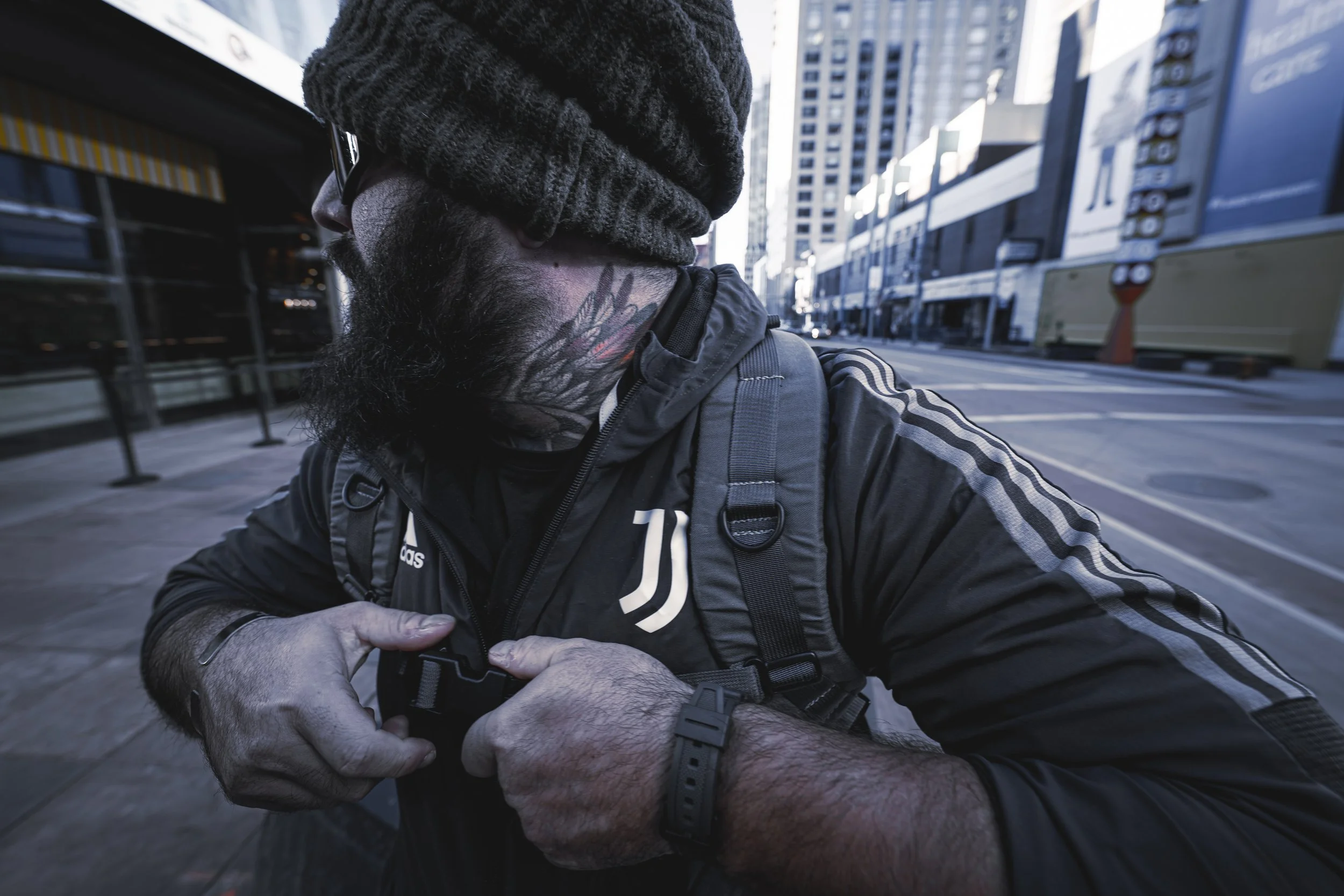 A bearded man wearing glasses, a black beanie, a black jacket with white stripes, and a backpack, adjusting his jacket strap on a city street with tall buildings in the background.