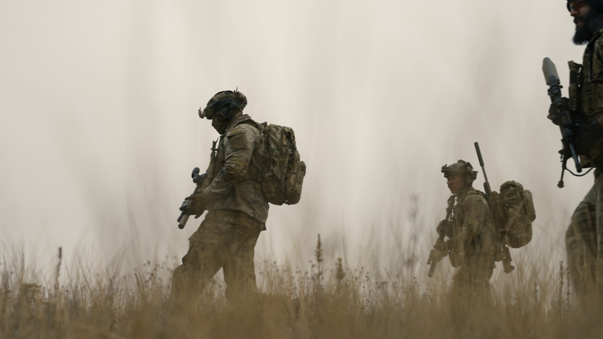 Three soldiers in camouflage gear walking through a grassy field with smoke around them.