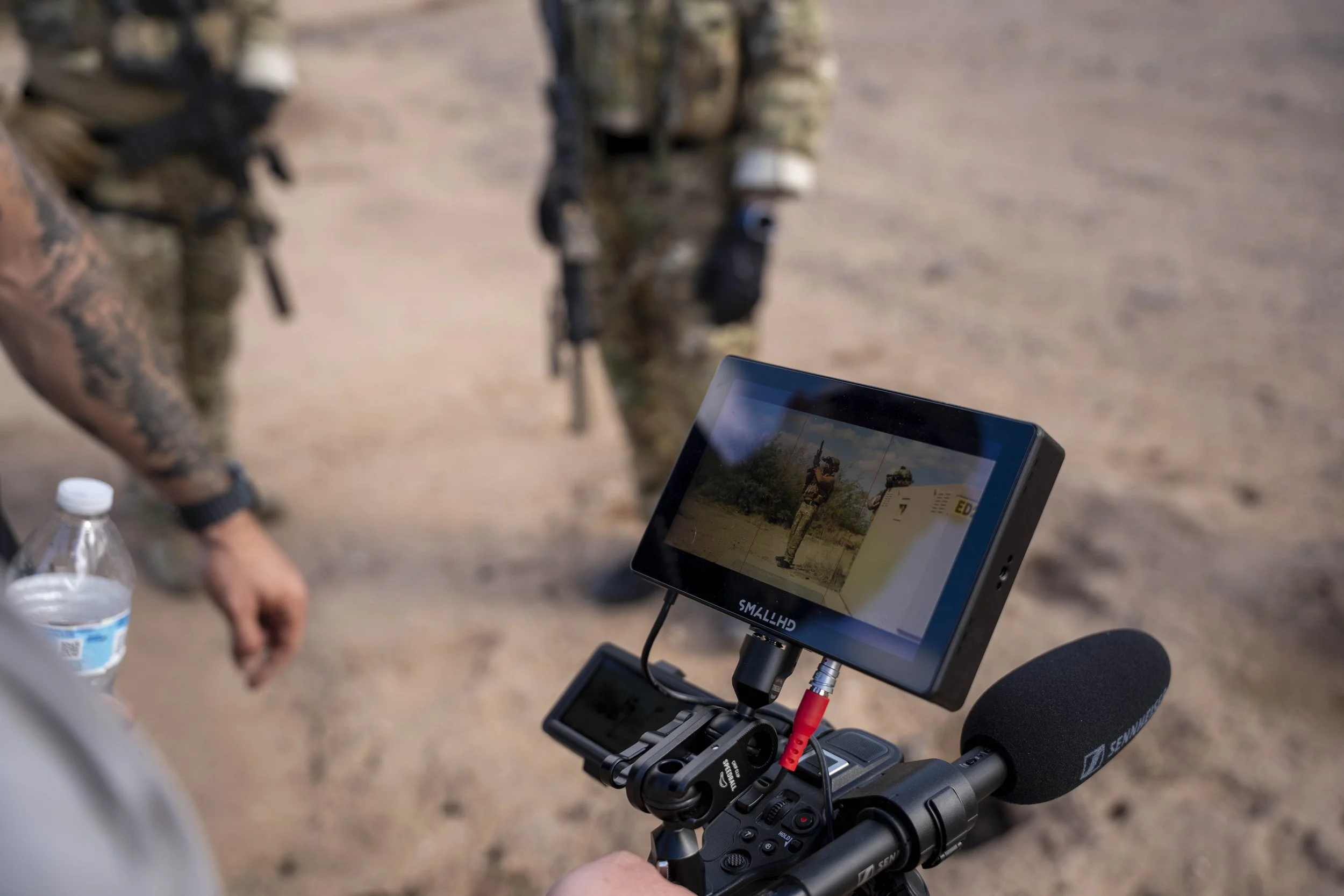 A camera mounted on a tripod with a monitor displaying an image of a soldier pointing at a wall, and a microphone attached, in a desert setting with soldiers in the background.