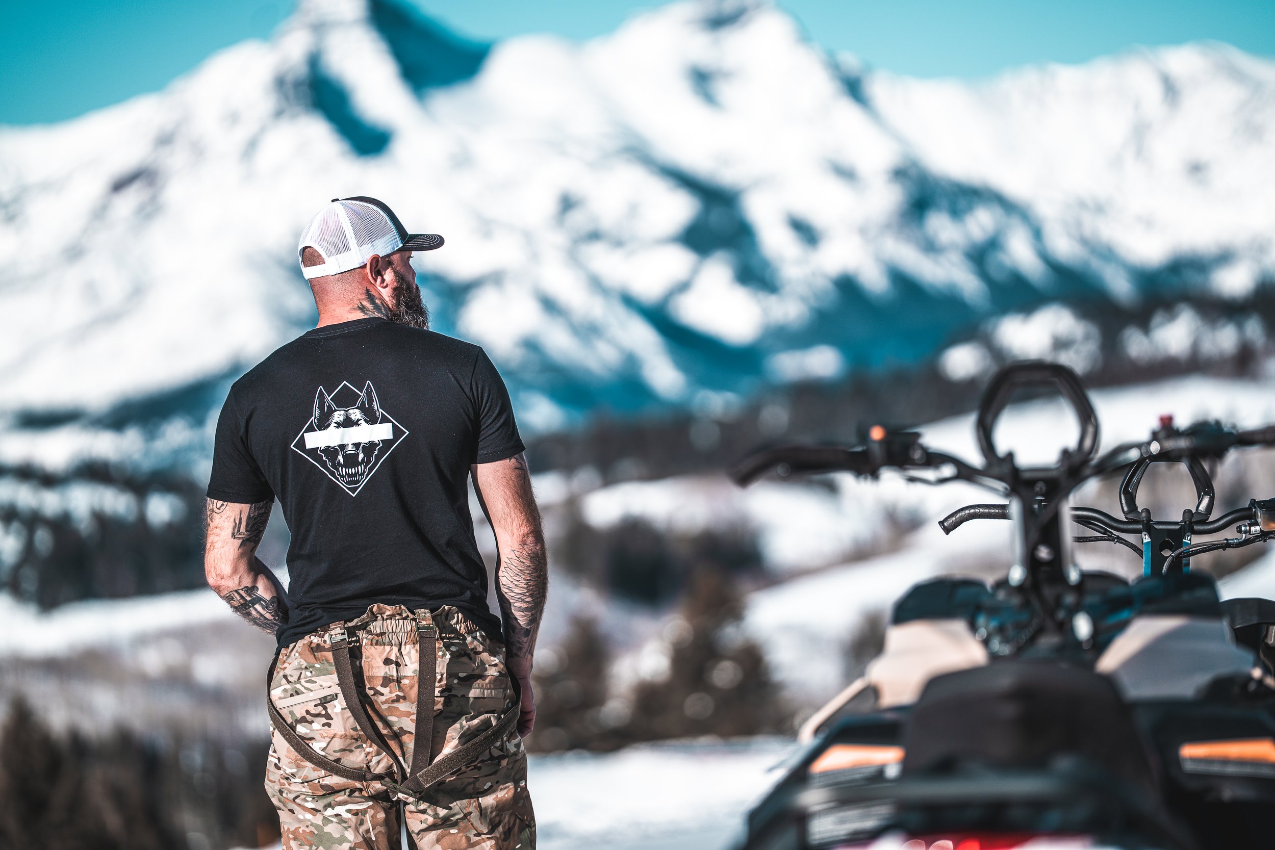A man standing outdoors in a snowy mountainous landscape, wearing a black t-shirt with a wolf emblem on the back, camouflage pants, and a white baseball cap, with snowmobiles nearby.