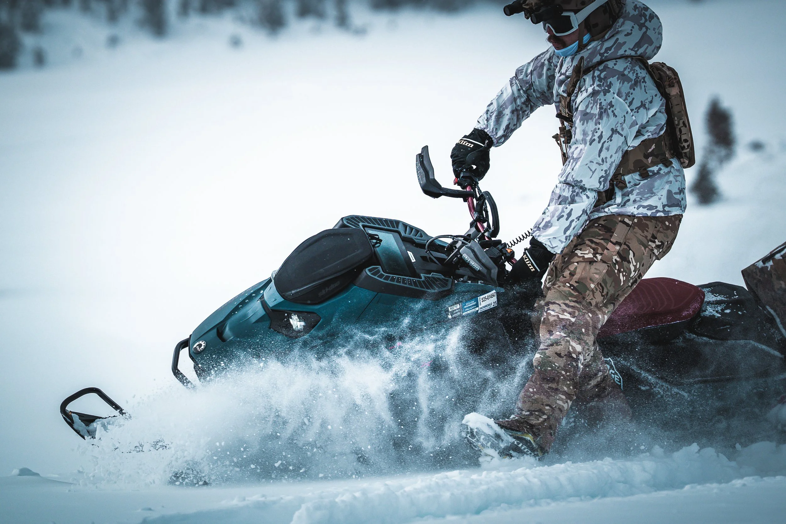 A person in camouflage winter gear riding a snowmobile through snow.