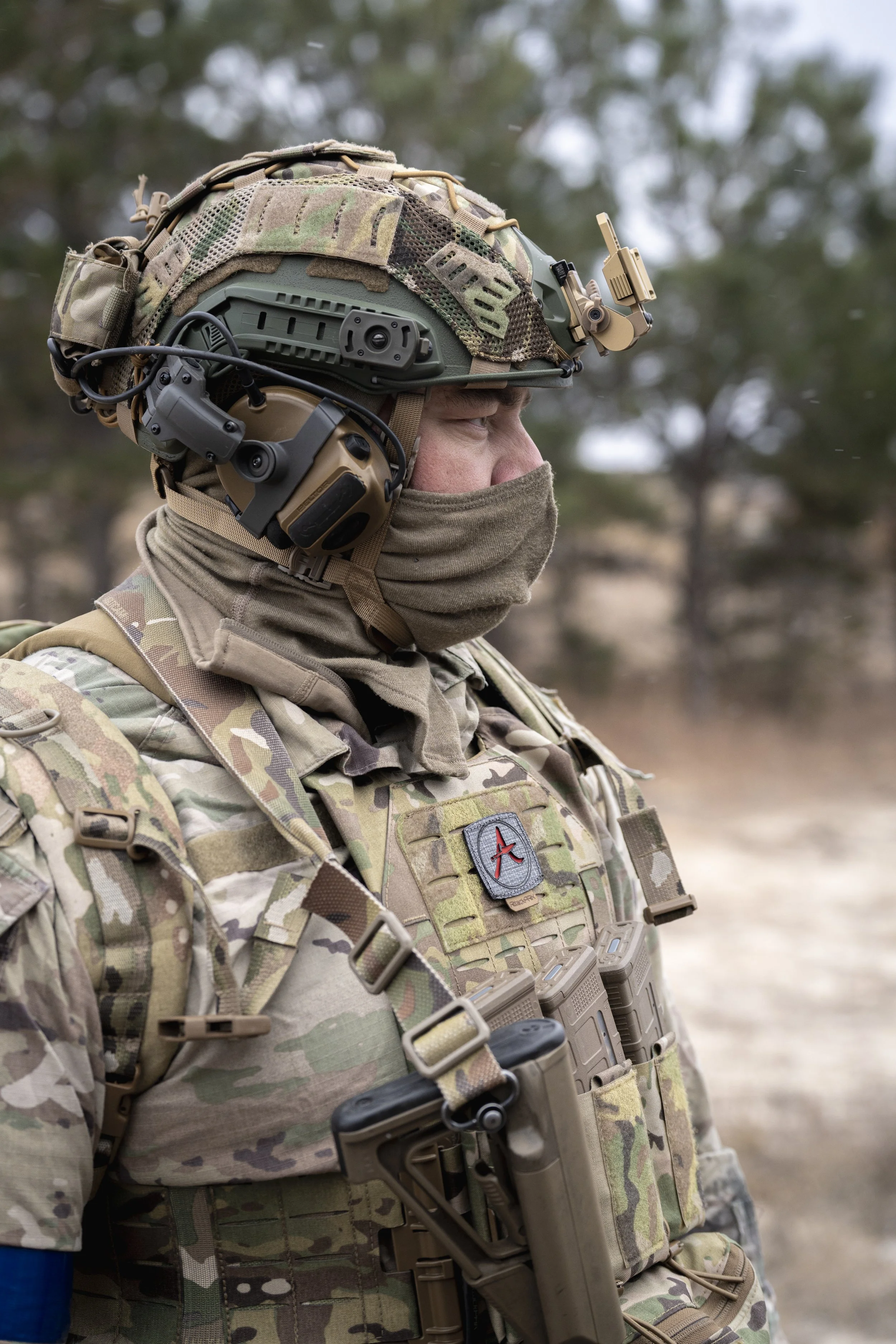 A soldier in full camouflage gear wearing tactical headphones, helmet, and face covering outside in a wooded area.
