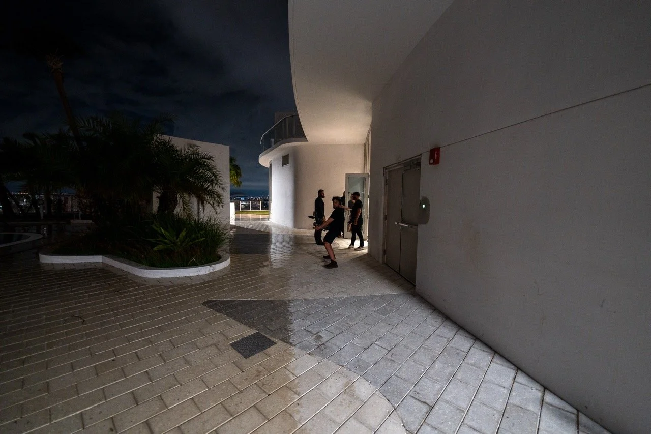 Nighttime scene on a rooftop terrace with four people standing near an elevator, some planting beds with plants, and an open view of the city in the distance.