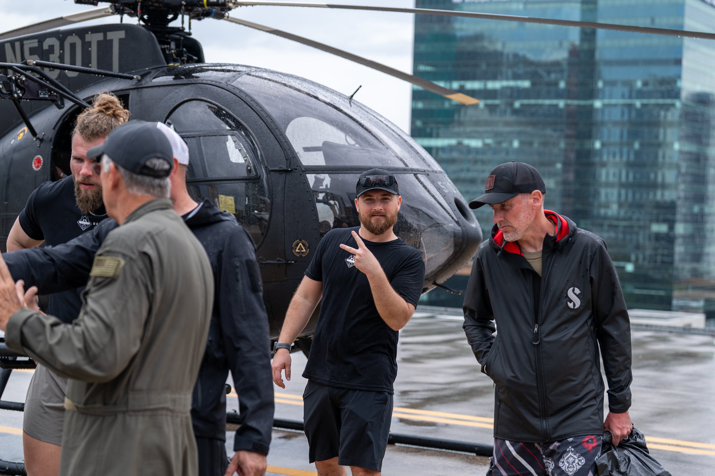 Group of men standing near a black helicopter on a rooftop with skyscrapers in the background, some wearing casual clothes and caps, rain appears to be falling.