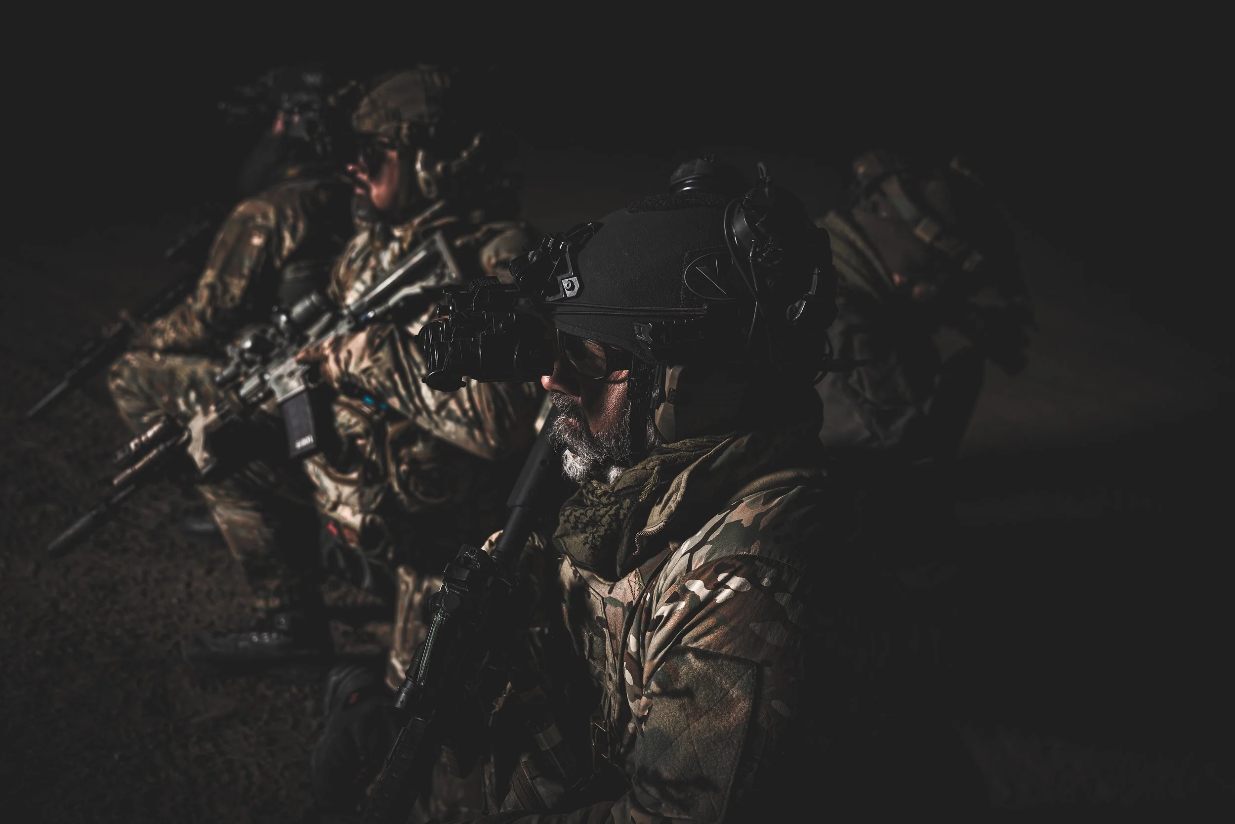 Military soldiers in camouflage gear and helmets, equipped with night vision goggles, sitting in a dark environment.