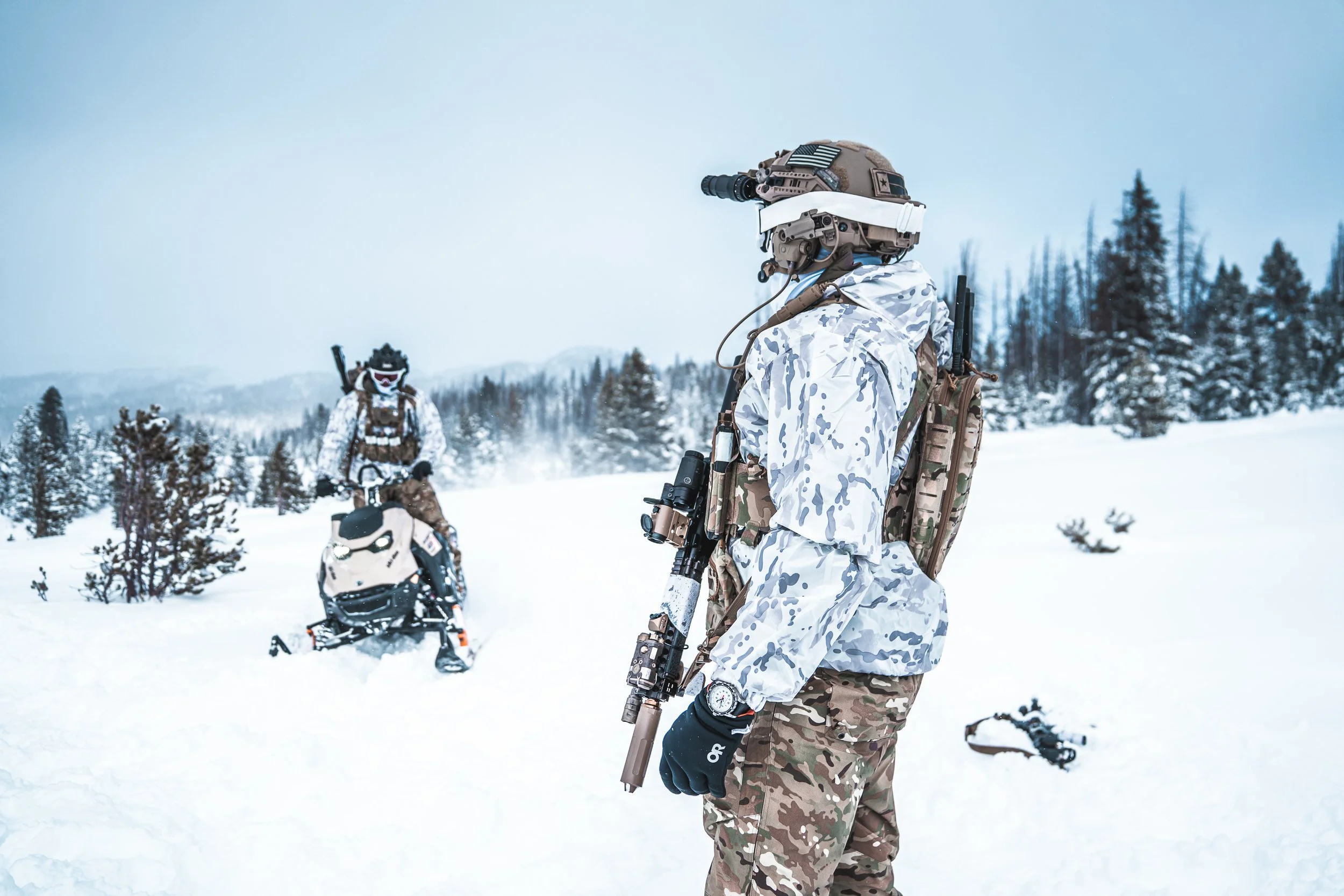 Two soldiers in camouflage winter gear with helmets and tactical gear in a snowy landscape, one standing and the other on a snowmobile.
