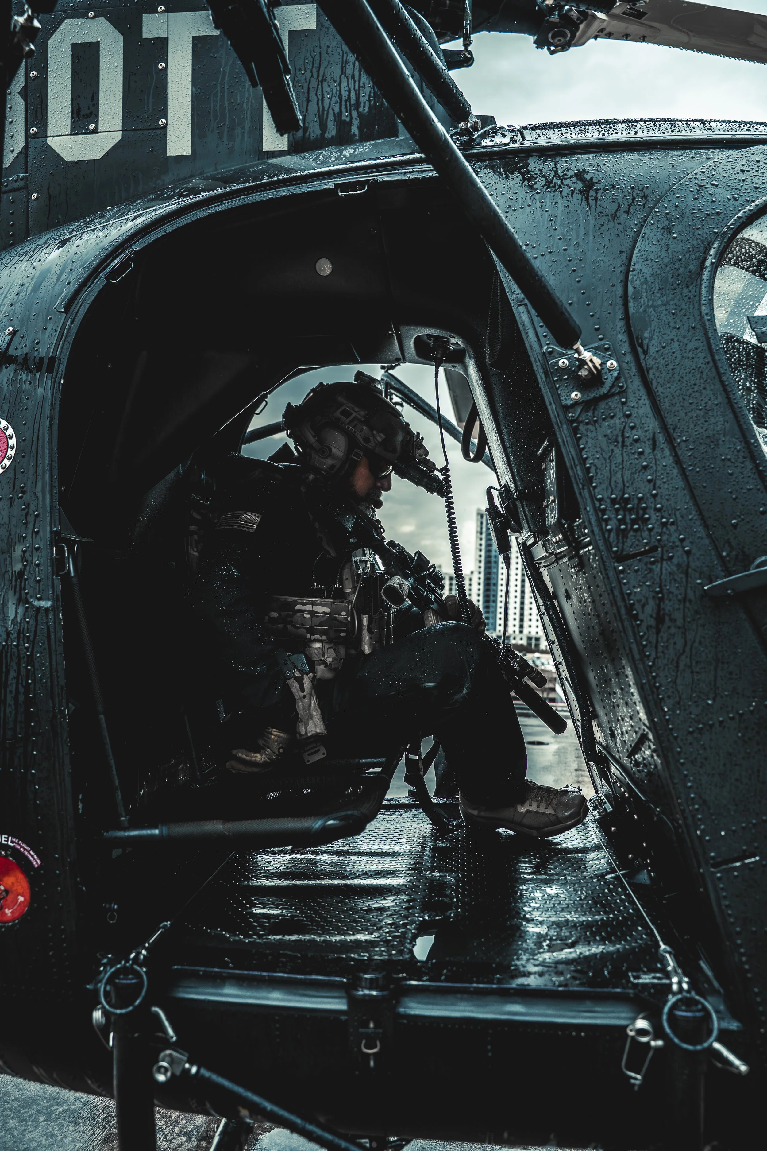 A soldier sitting inside a black helicopter cockpit, wearing tactical gear and a helmet, on a rainy day.