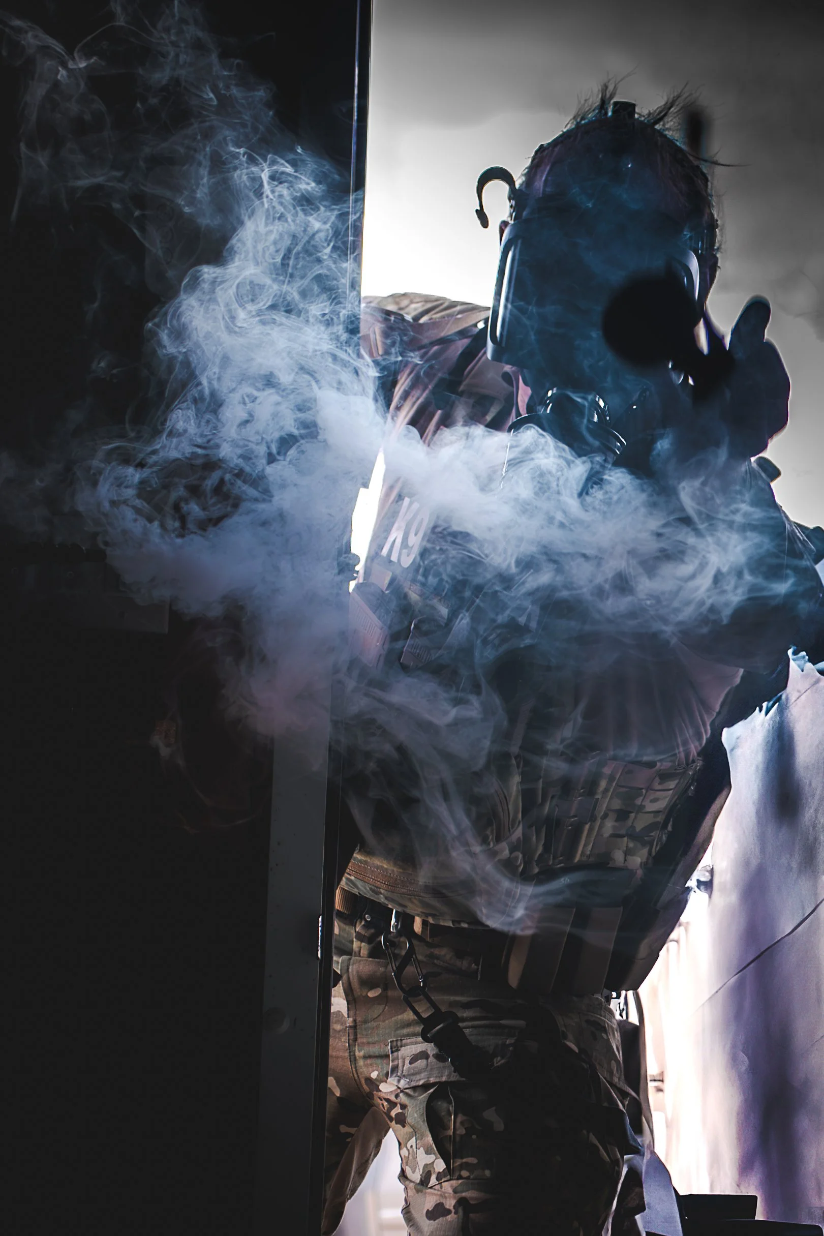 A person wearing tactical gear and a gas mask is partially obscured by smoke, looking at a computer screen or device in a dark environment.