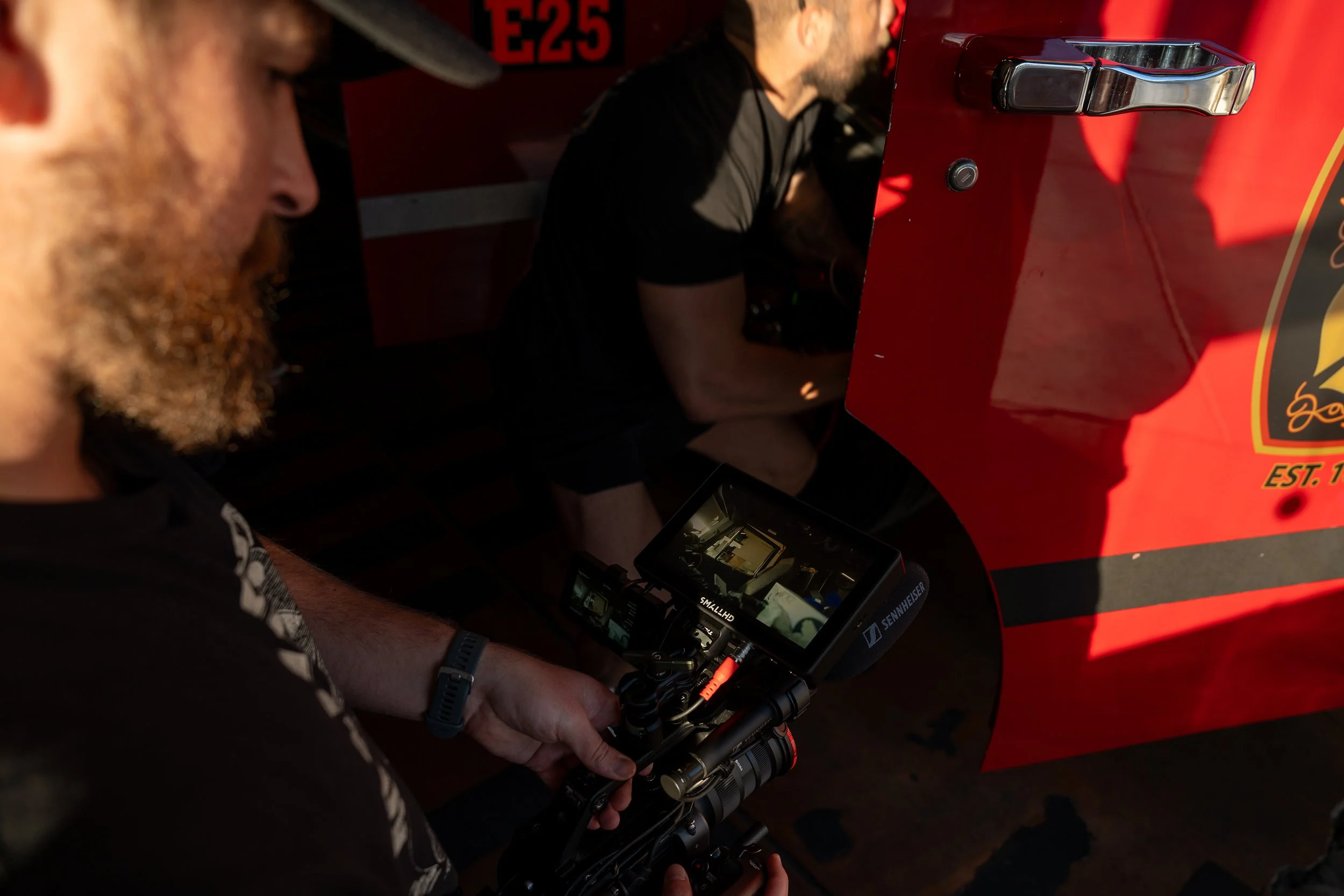 Two men are working with a camera and a monitor next to a red fire truck. One man with a beard and dark clothing is using the camera, and the other man is leaning over the fire truck.