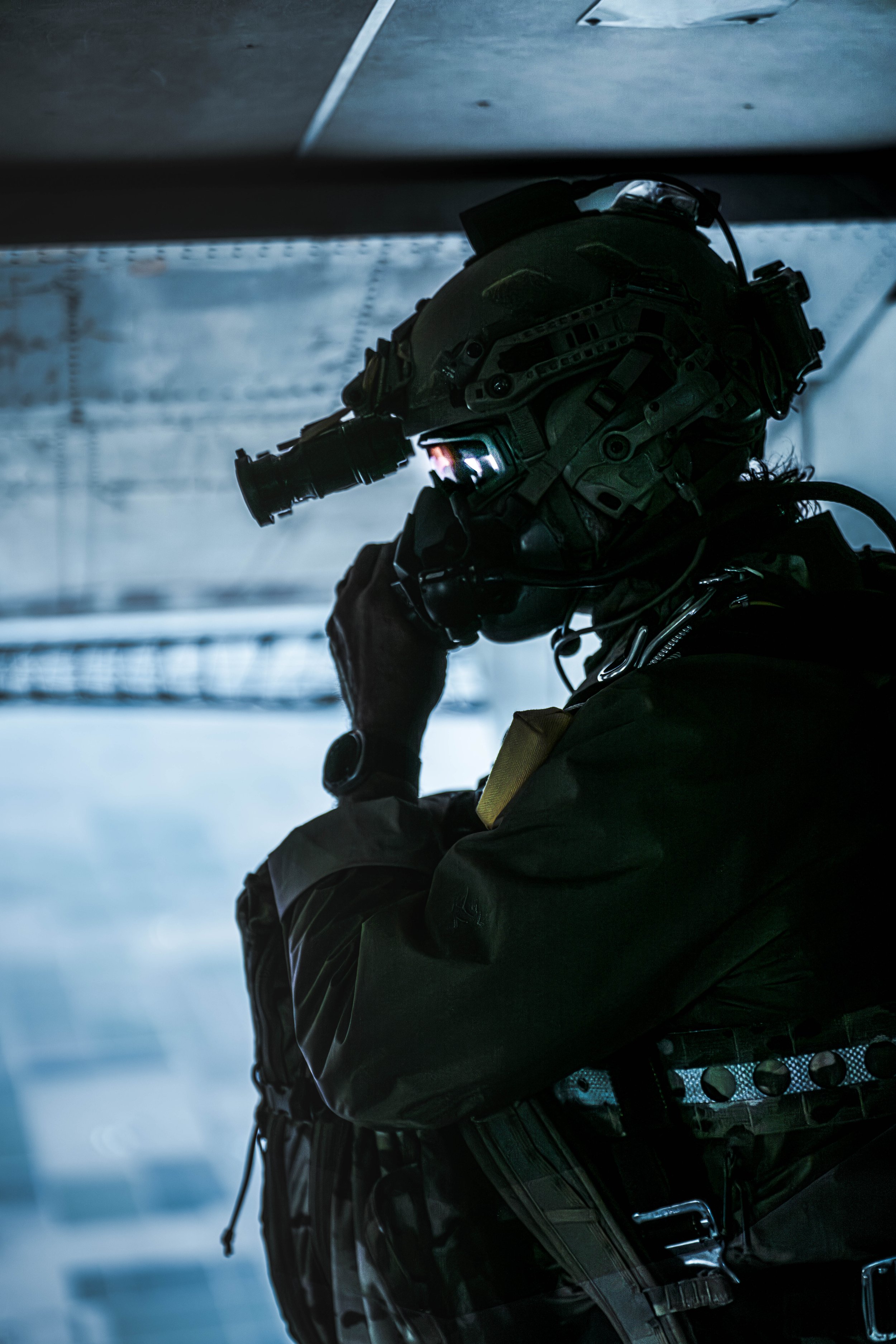 A military personnel wearing tactical gear and a helmet with night vision goggles, observing from a shadowed area.