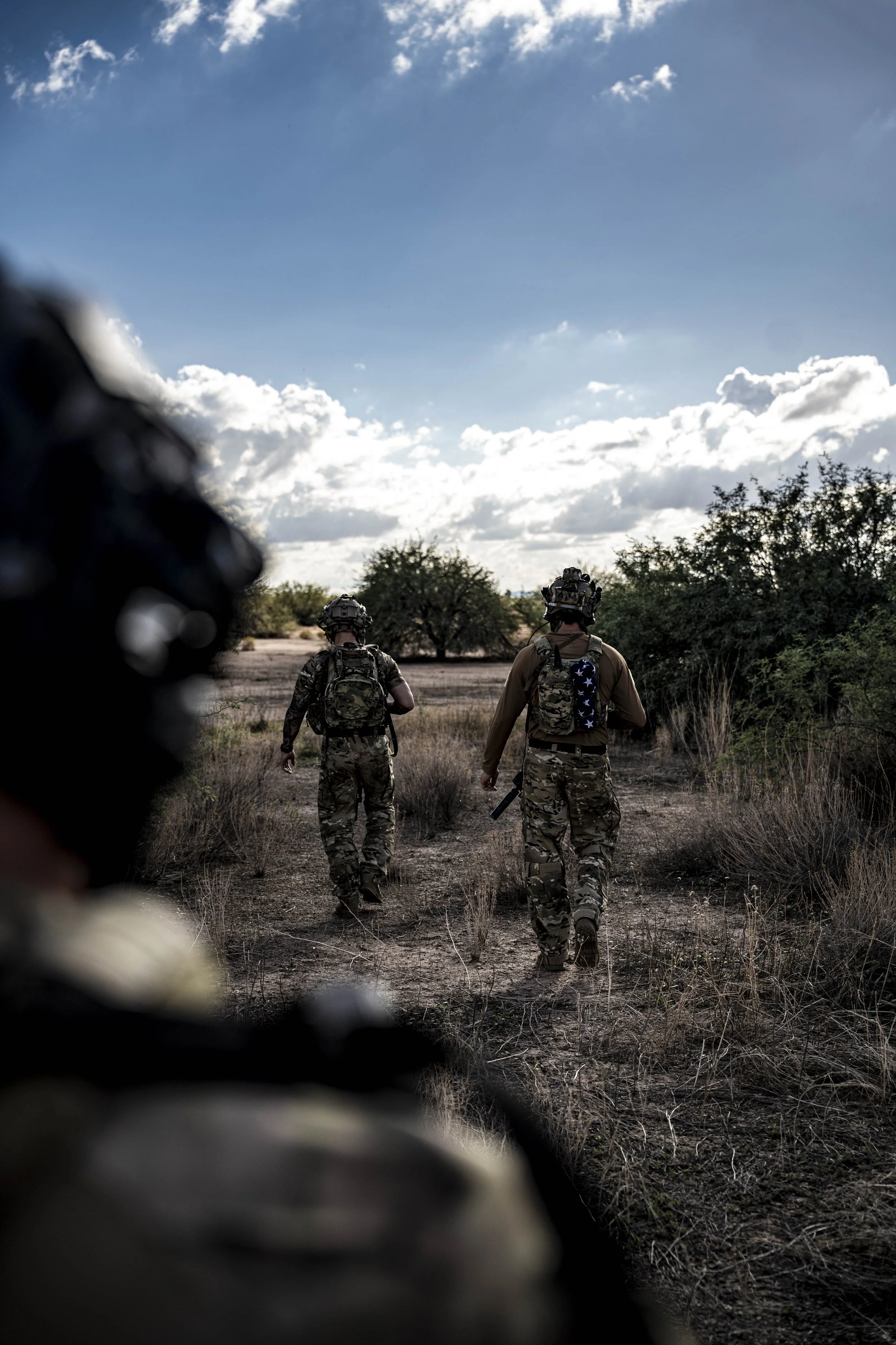 Three soldiers walking in a dry, grassy landscape with trees, under a partly cloudy sky, with a blurred object in the foreground.