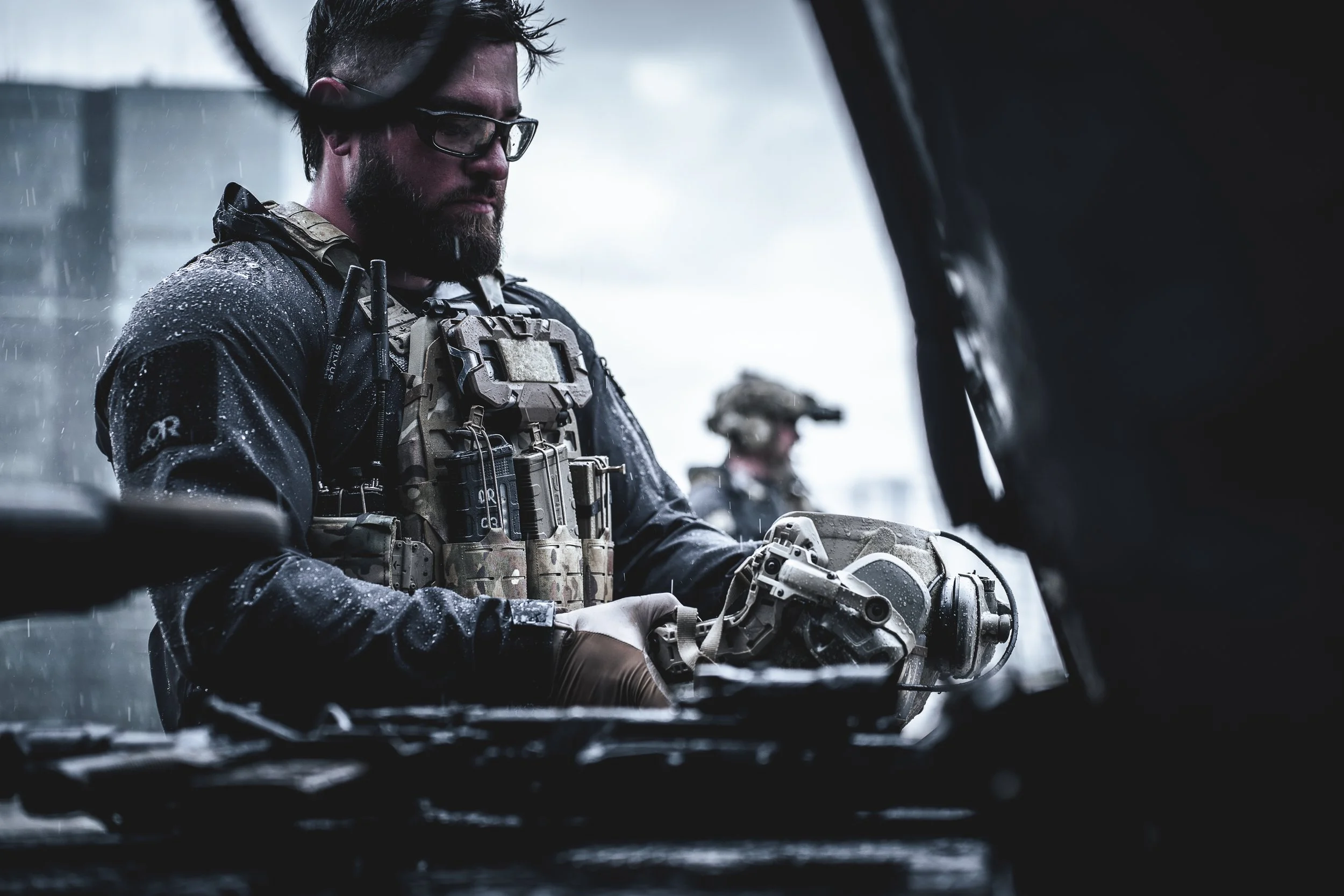 Military personnel working on equipment in the rain, wearing tactical gear and glasses.