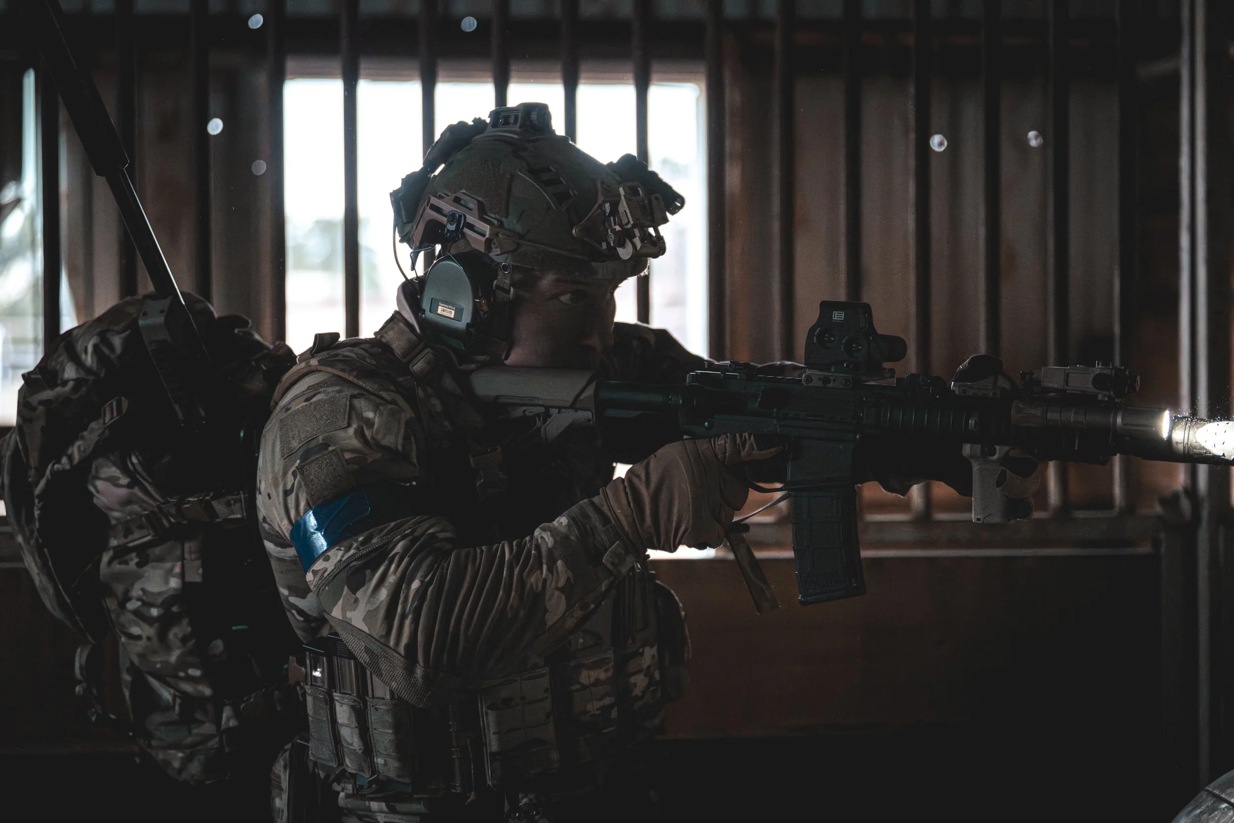 A soldier in camouflage uniform aiming a rifle with attached sight inside a wooden building with barred windows.