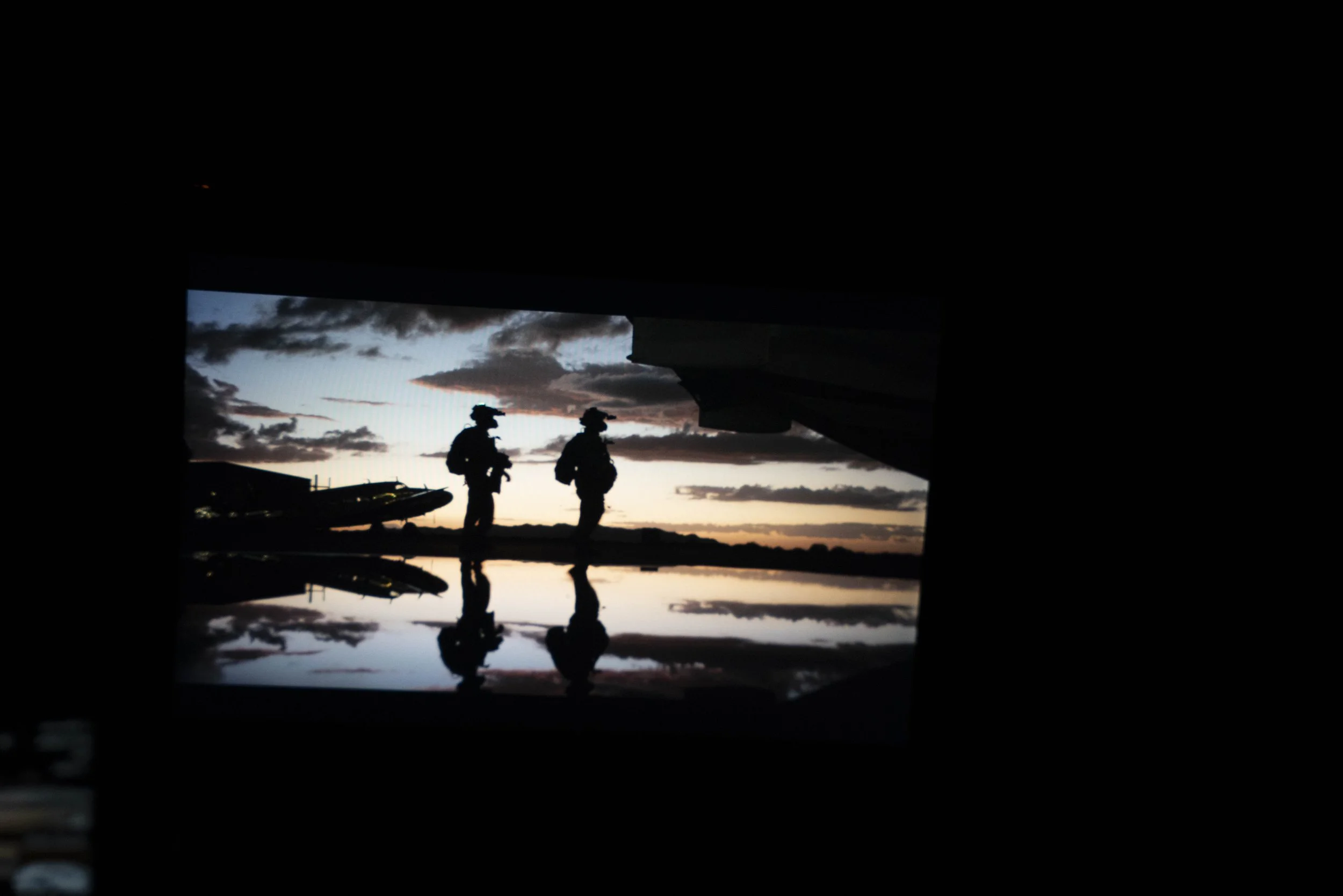 Silhouettes of two soldiers walking outside during sunset, with aircraft and clouds in the background, reflected on a wet surface.