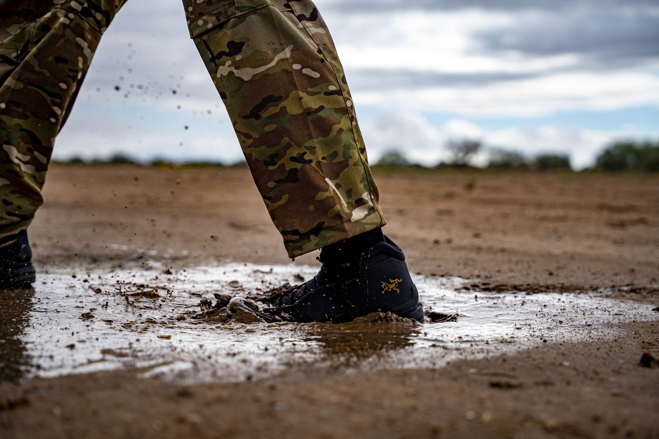 Close-up of person in camouflage pants and black outdoor shoes splashing through wet mud on a dirt field with a cloudy sky in the background.