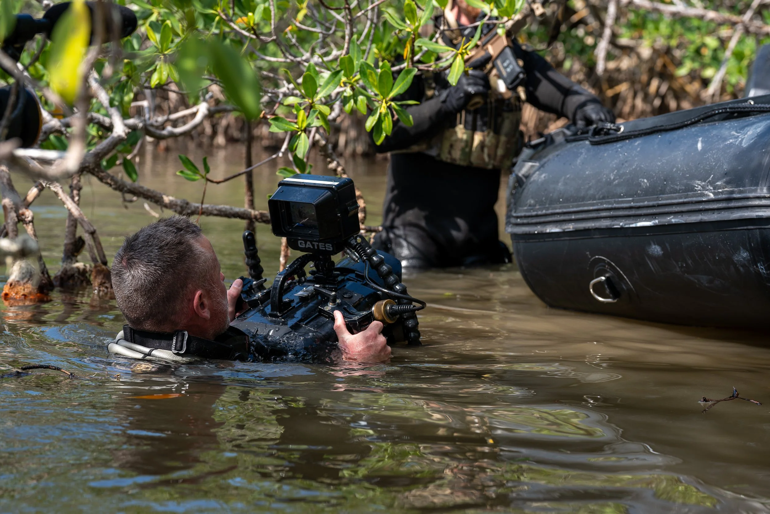 A person in a wetsuit with a camera in a watery environment, possibly a flood or a river, with another person partially visible in the background near some vegetation.