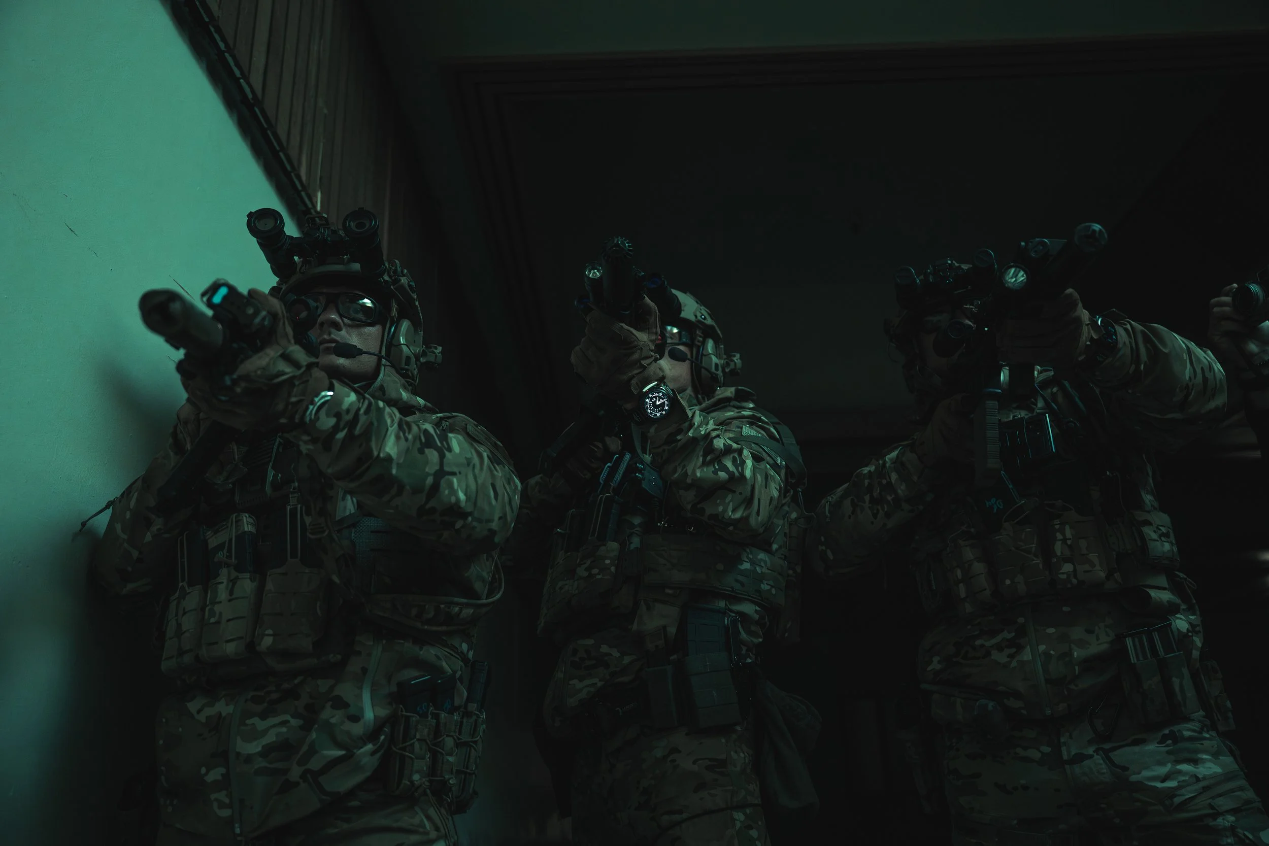 Military personnel in camouflage gear aiming rifles in a dark indoor setting.