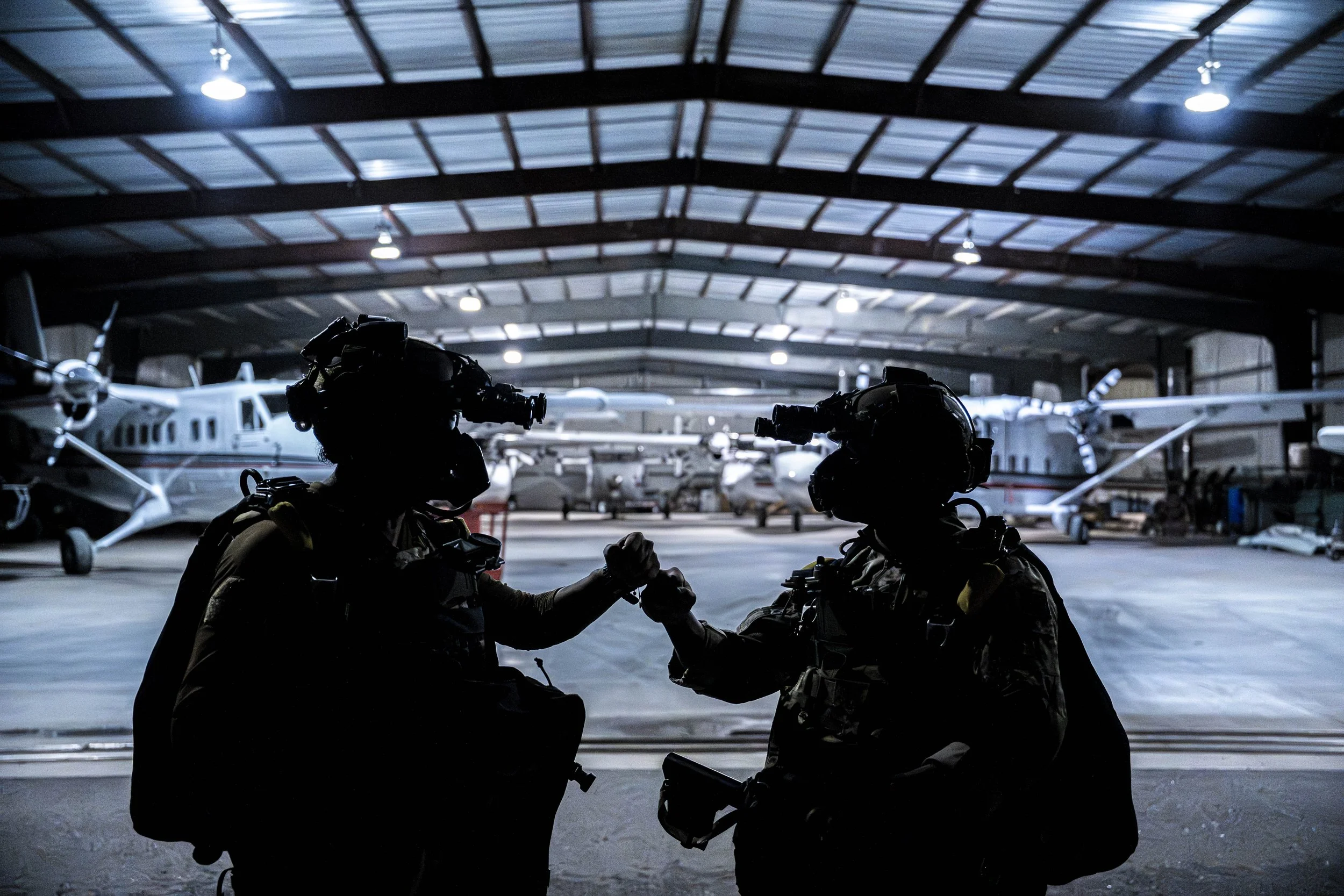 Silhouettes of two soldiers in tactical gear shaking hands in an aircraft hangar with planes in the background.