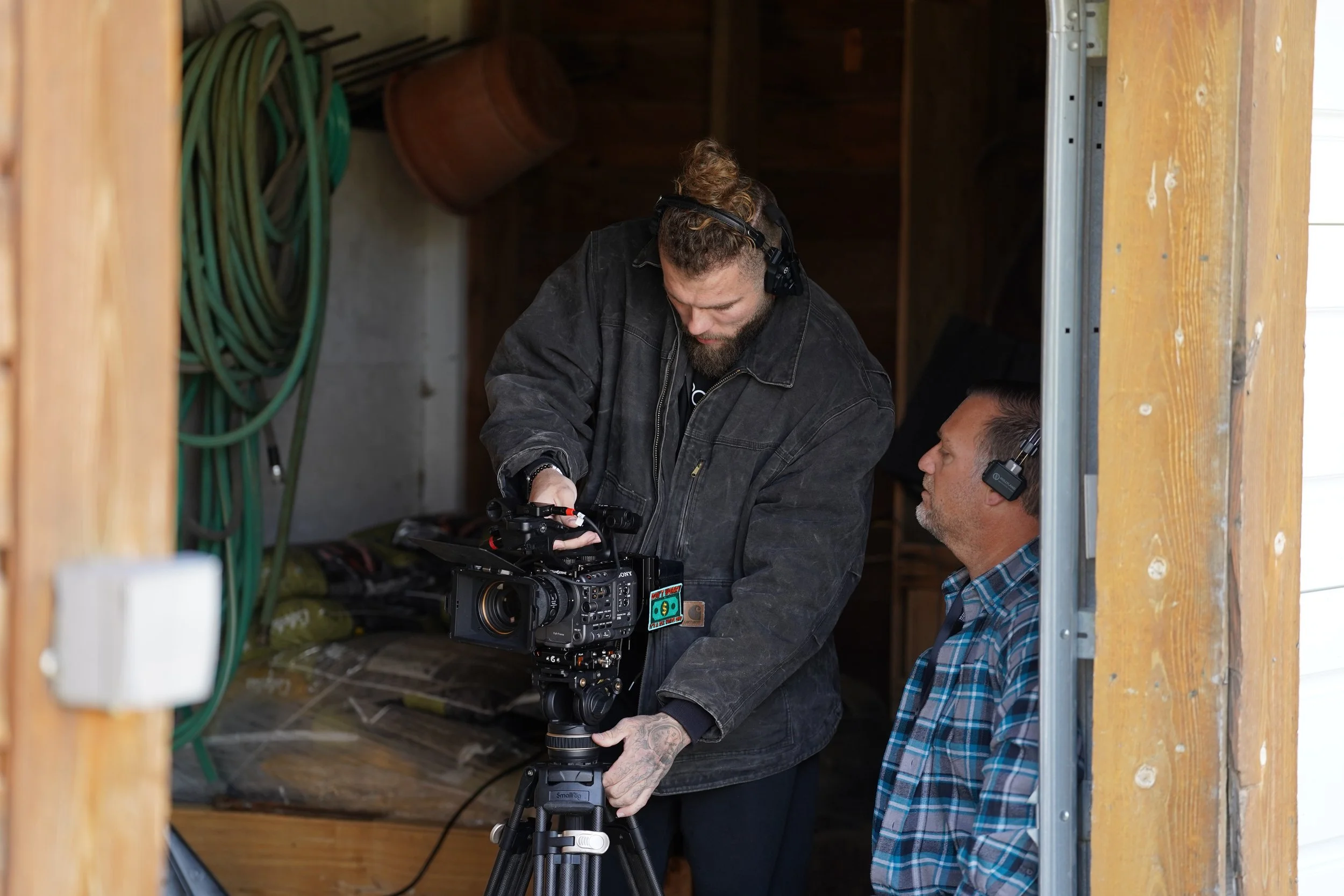 Two men working with professional video camera equipment inside a wooden shed, one man is standing and adjusting the camera, the other man is sitting and observing, both wearing headsets.
