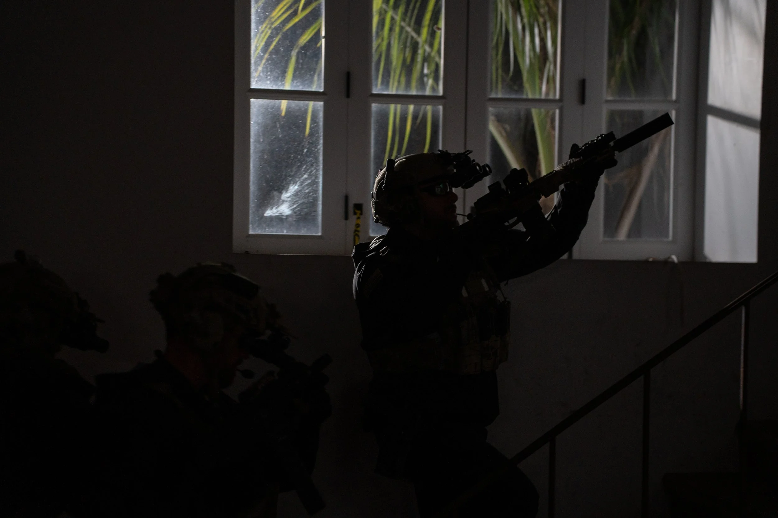 Silhouetted soldiers with rifles inside a dark room near a window.