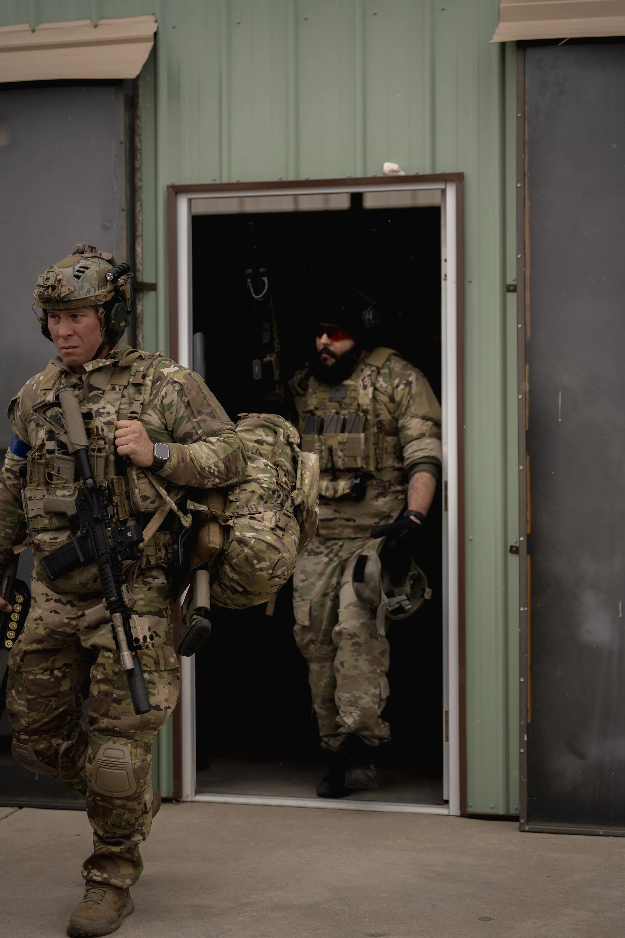 Two soldiers in camouflage uniforms exiting a green building, one walking out with a backpack and a rifle, the other seated inside holding a helmet.