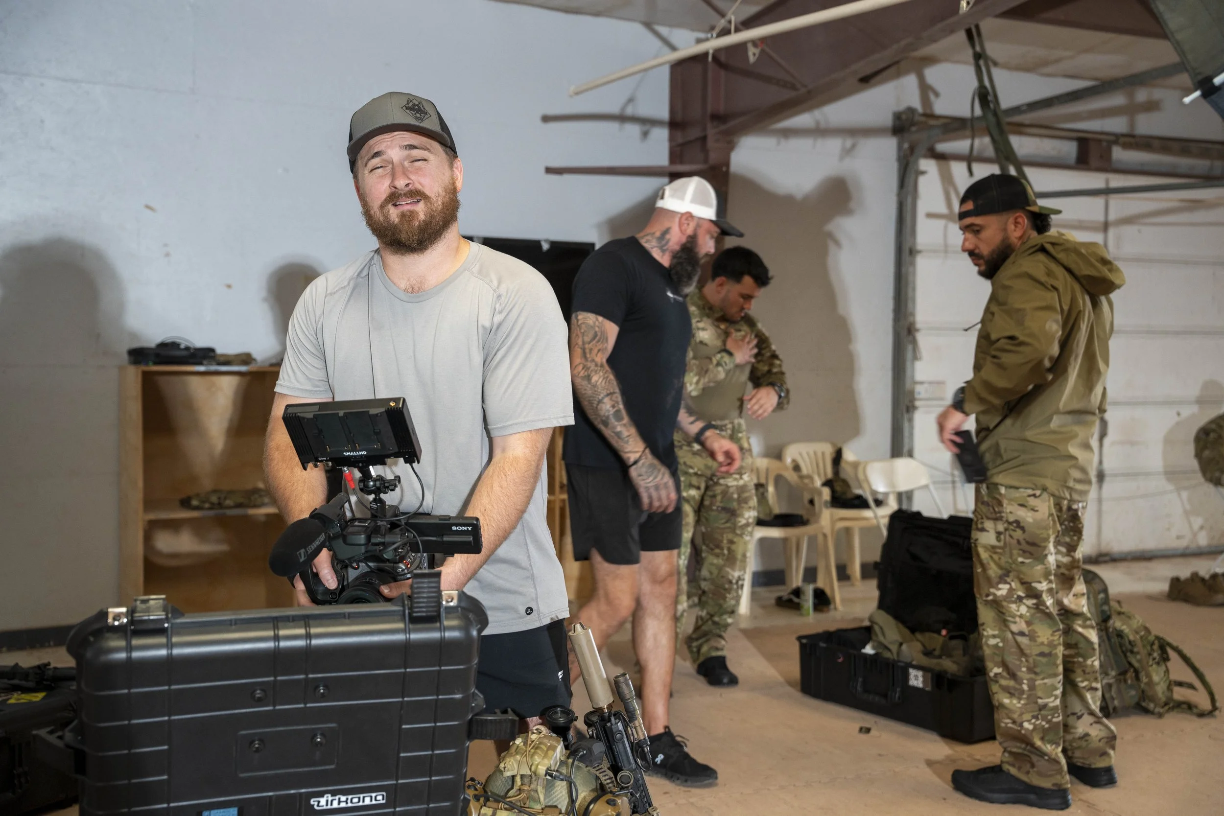 Group of four men in a warehouse with gear and equipment, one holding a camera, others preparing gear, all dressed casually or in military style clothing.