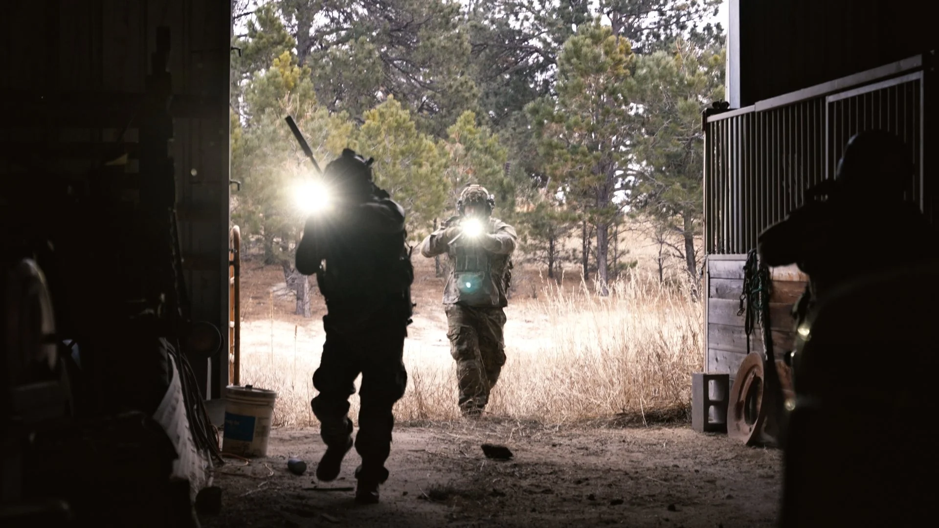 Three armed individuals exiting a barn or garage into a field with trees, with bright sunlight behind them.