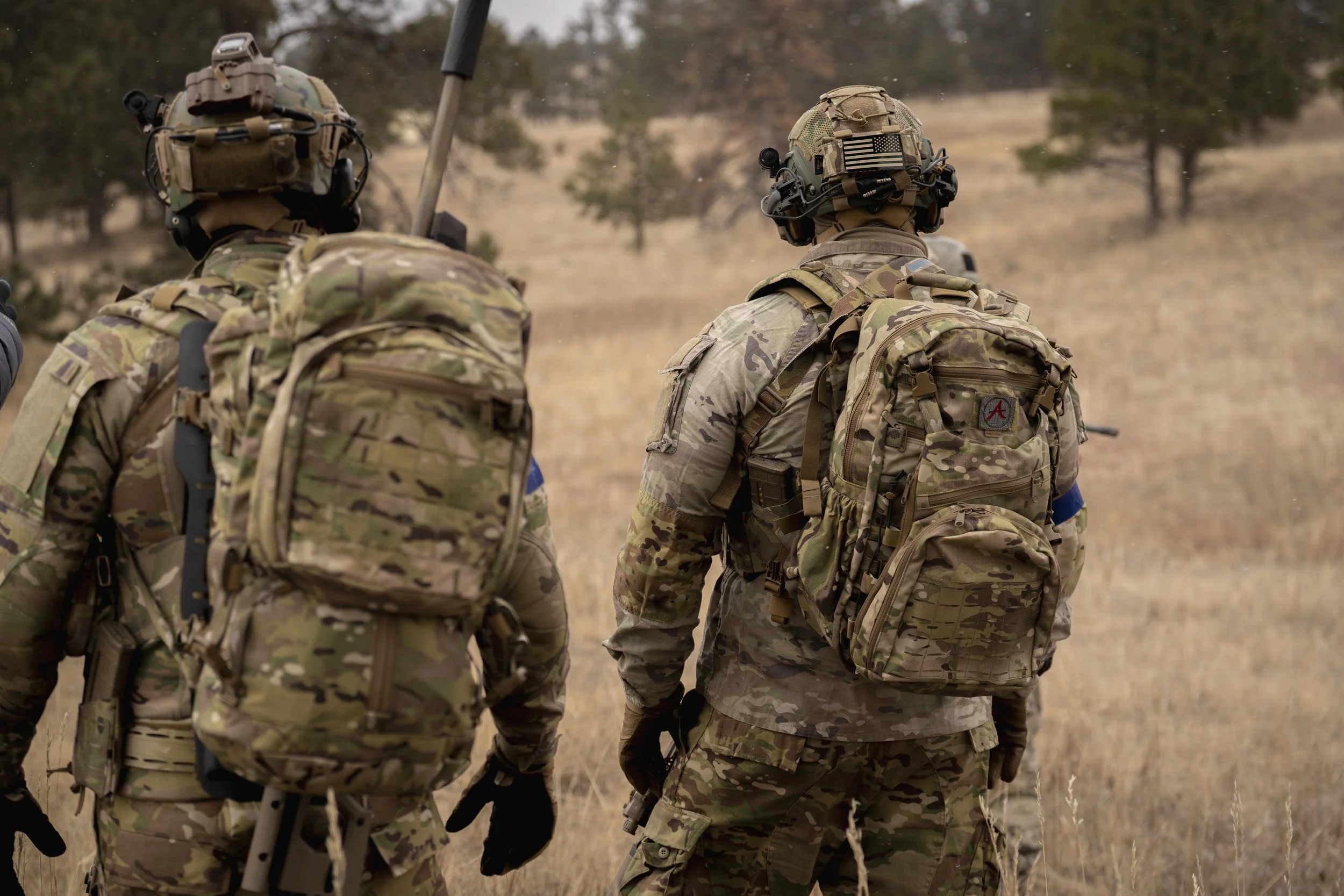 Three soldiers in camouflage uniforms walking through a dry grassy field with trees in the background, carrying backpacks and gear.