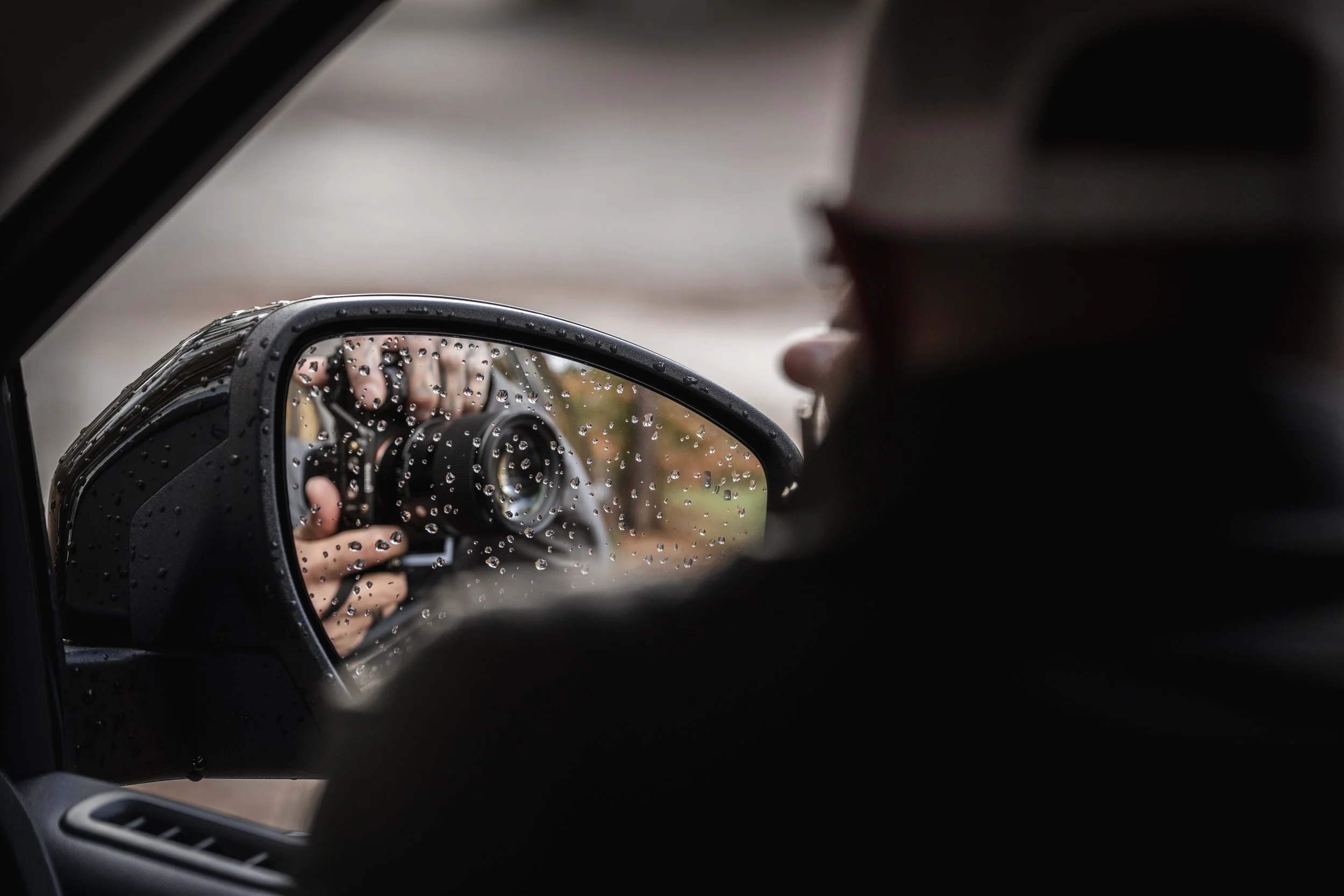 A person with a hat taking a photo of the view through a rainy car side mirror, which has raindrops on it.