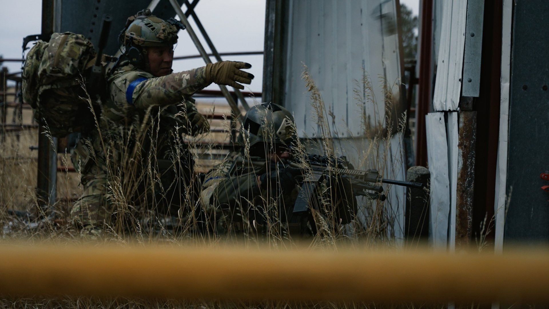 Two soldiers in camouflage uniforms and helmets taking cover behind a building, one aiming a rifle while the other gestures.