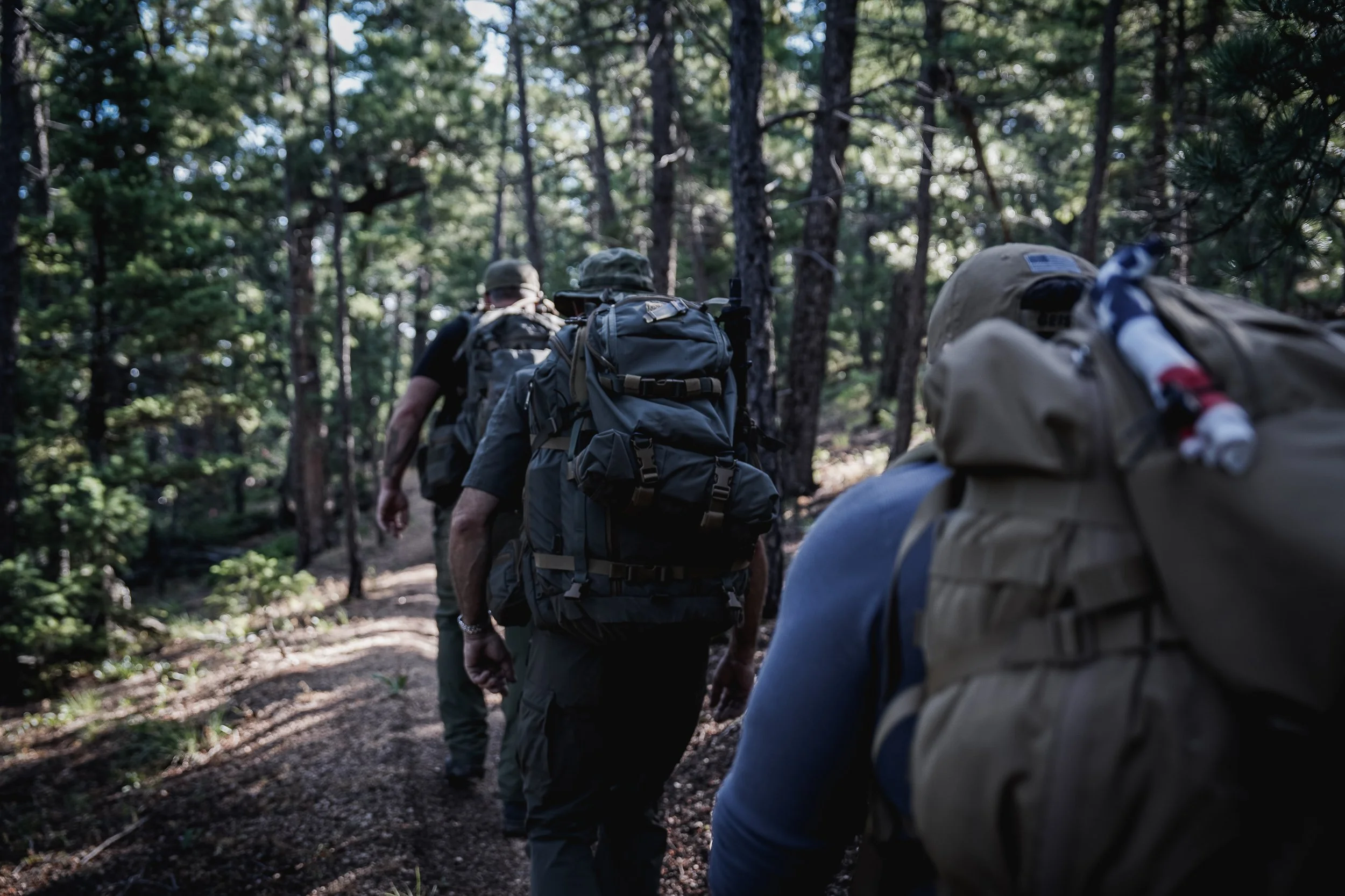Group of people hiking in a forest with backpacks on a dirt trail surrounded by tall trees.