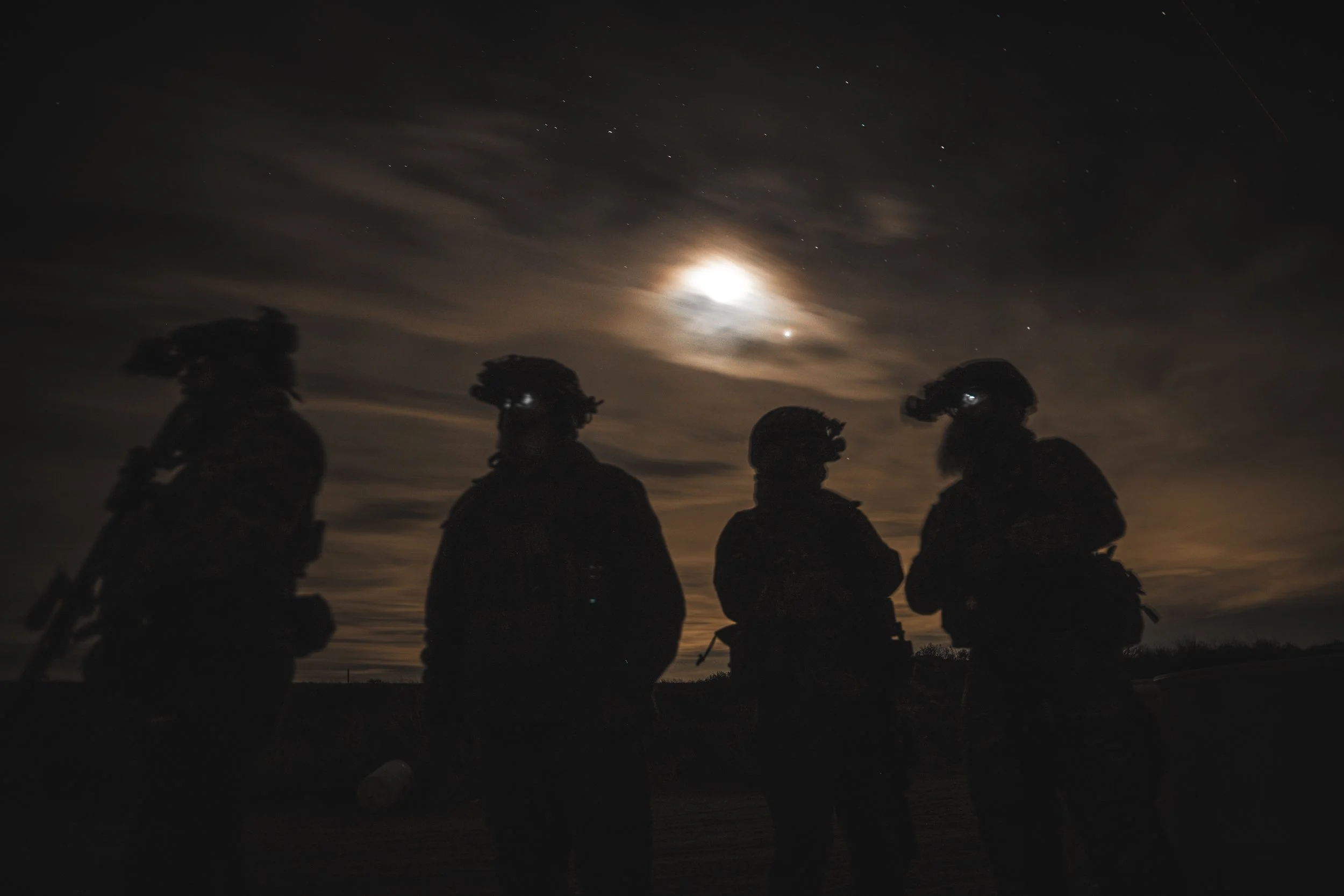 Four silhouetted soldiers standing outdoors at night with a moonlit sky and clouds in the background.