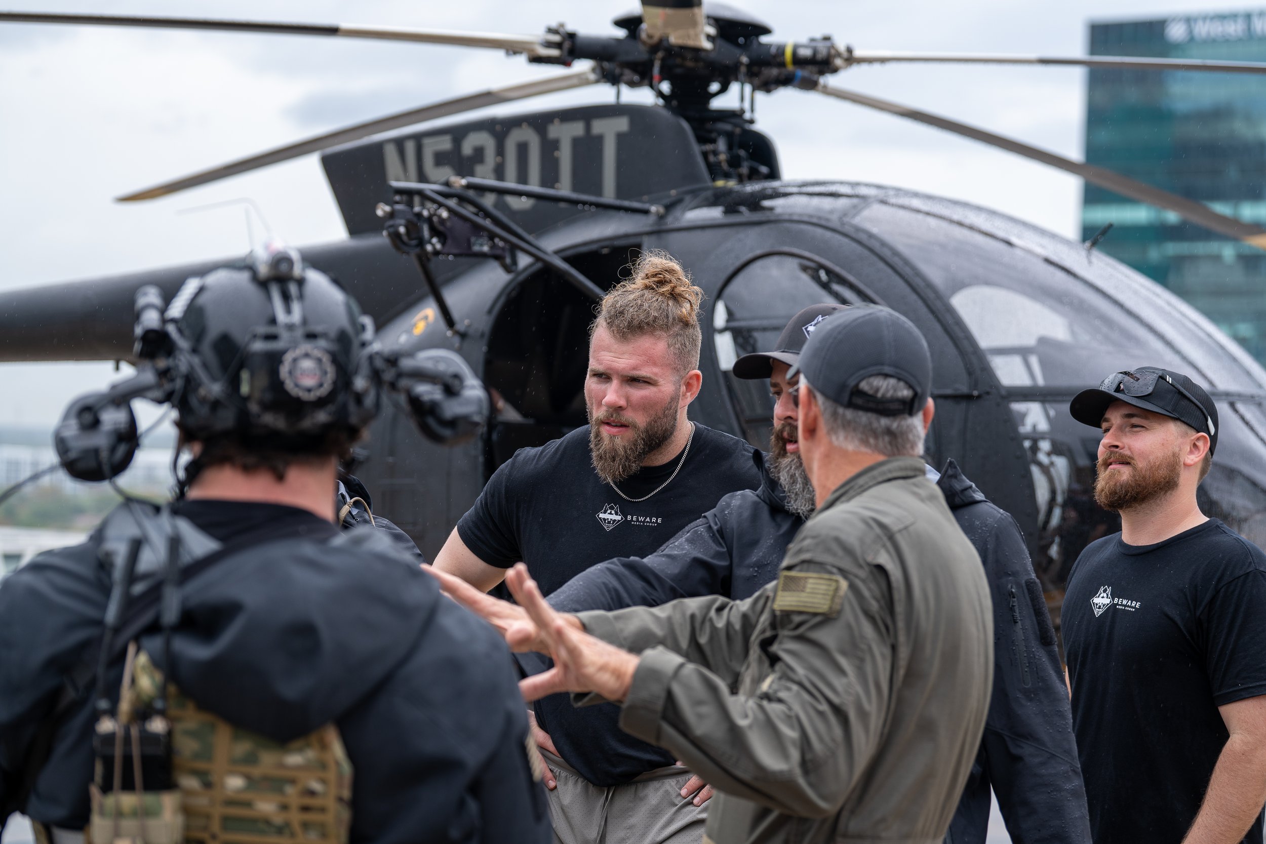 Group of military or special operations personnel, some with tactical gear, gathered around a man in a black t-shirt with a helicopter in the background.