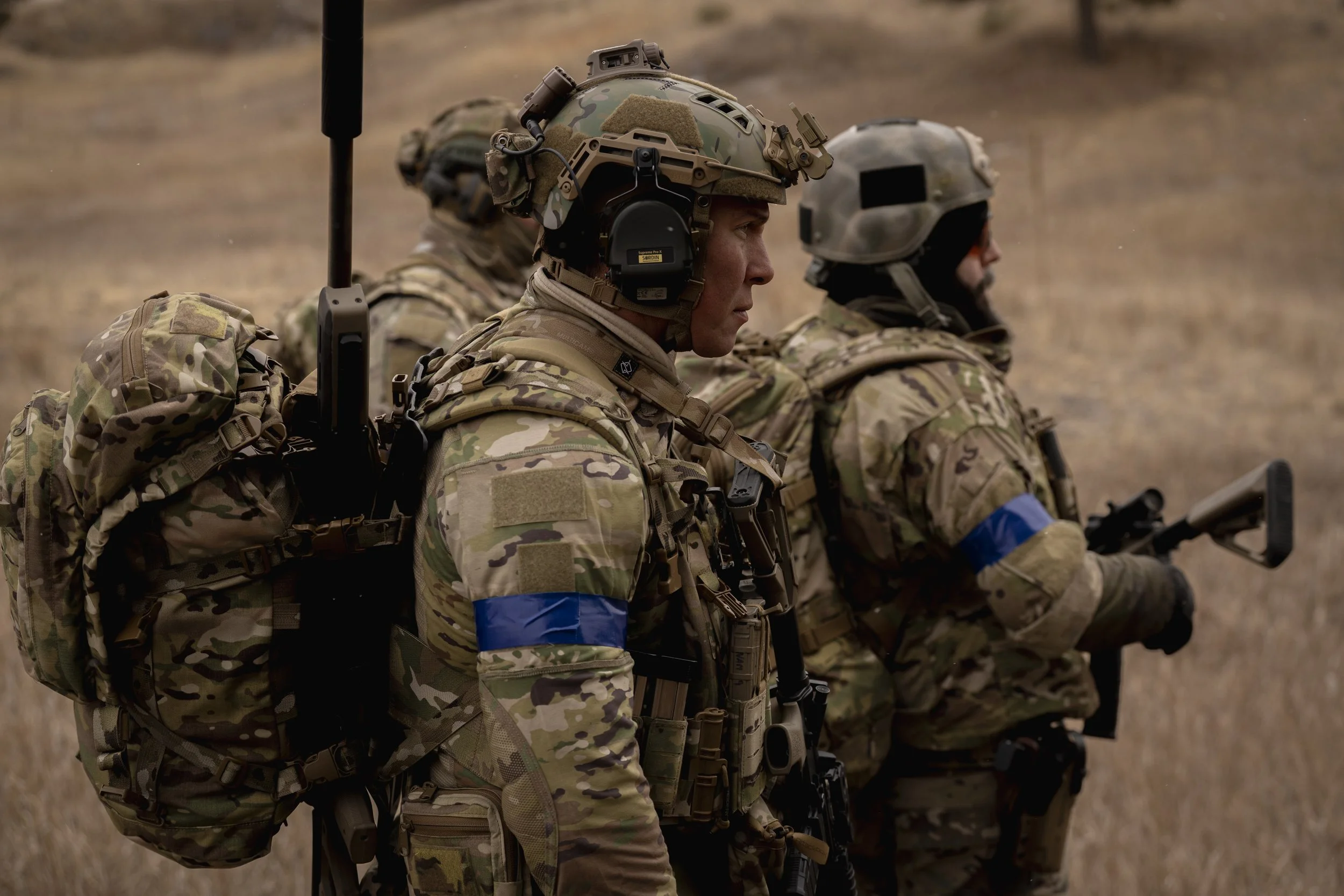 Army soldiers in camouflage uniform with gear, helmets, and blue arm bands standing in a field.
