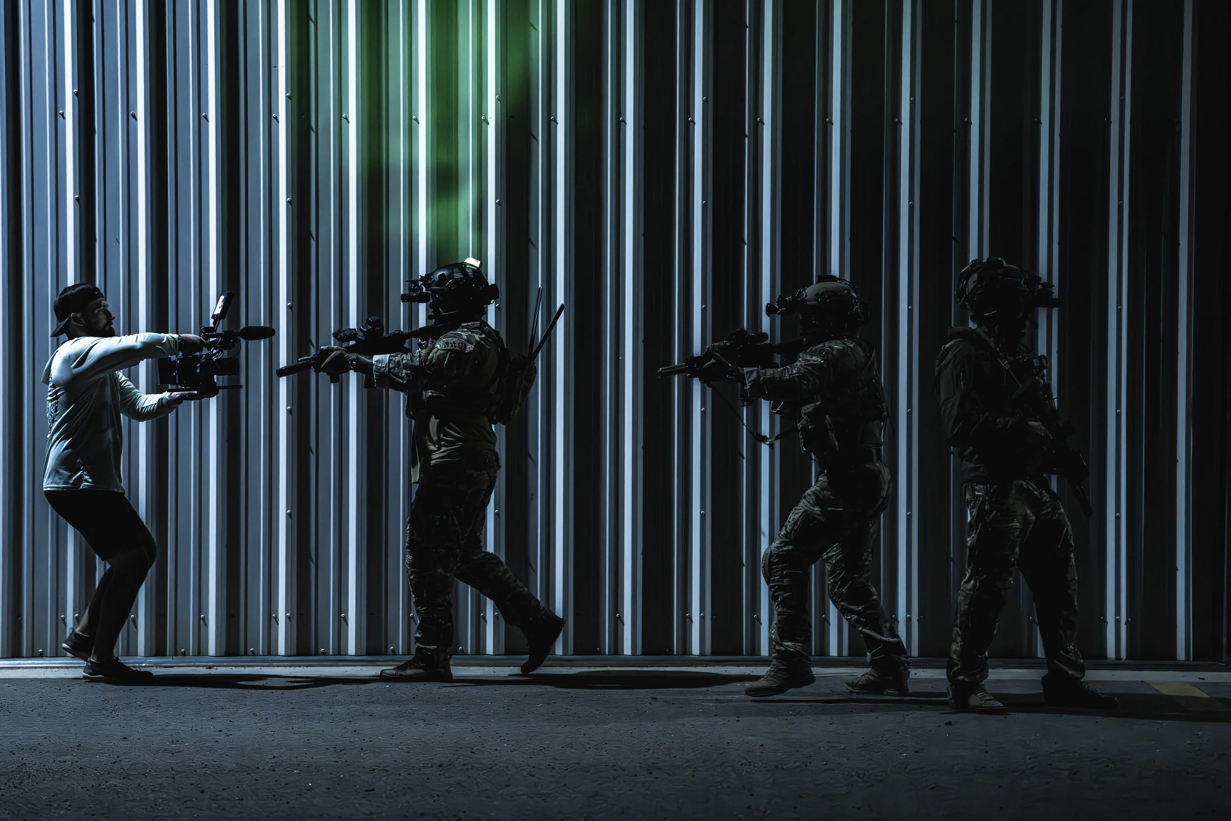 A person on the left appears to be taking a photograph of three armed soldiers in tactical gear against a corrugated metal wall at night.