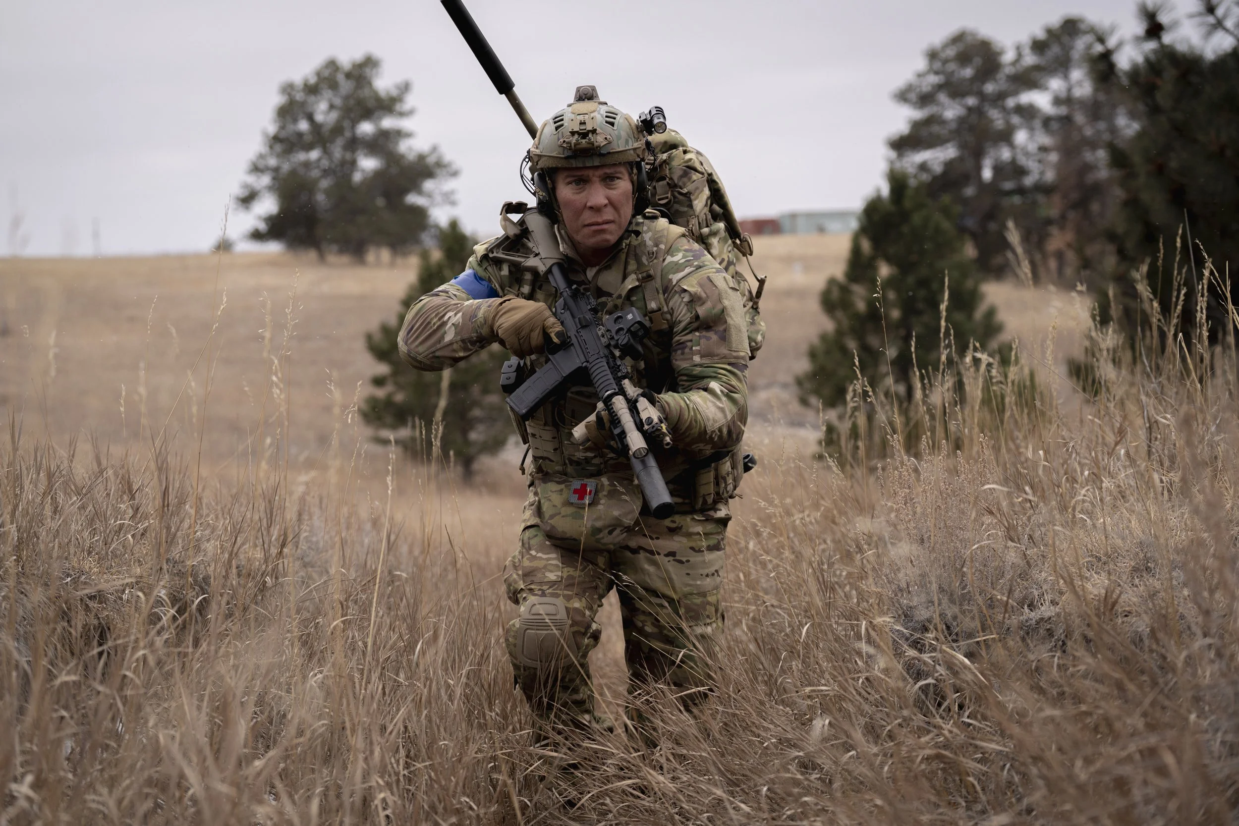 A soldier in camouflage uniform walking through a grassy field, holding a rifle, with trees and a building in the background.